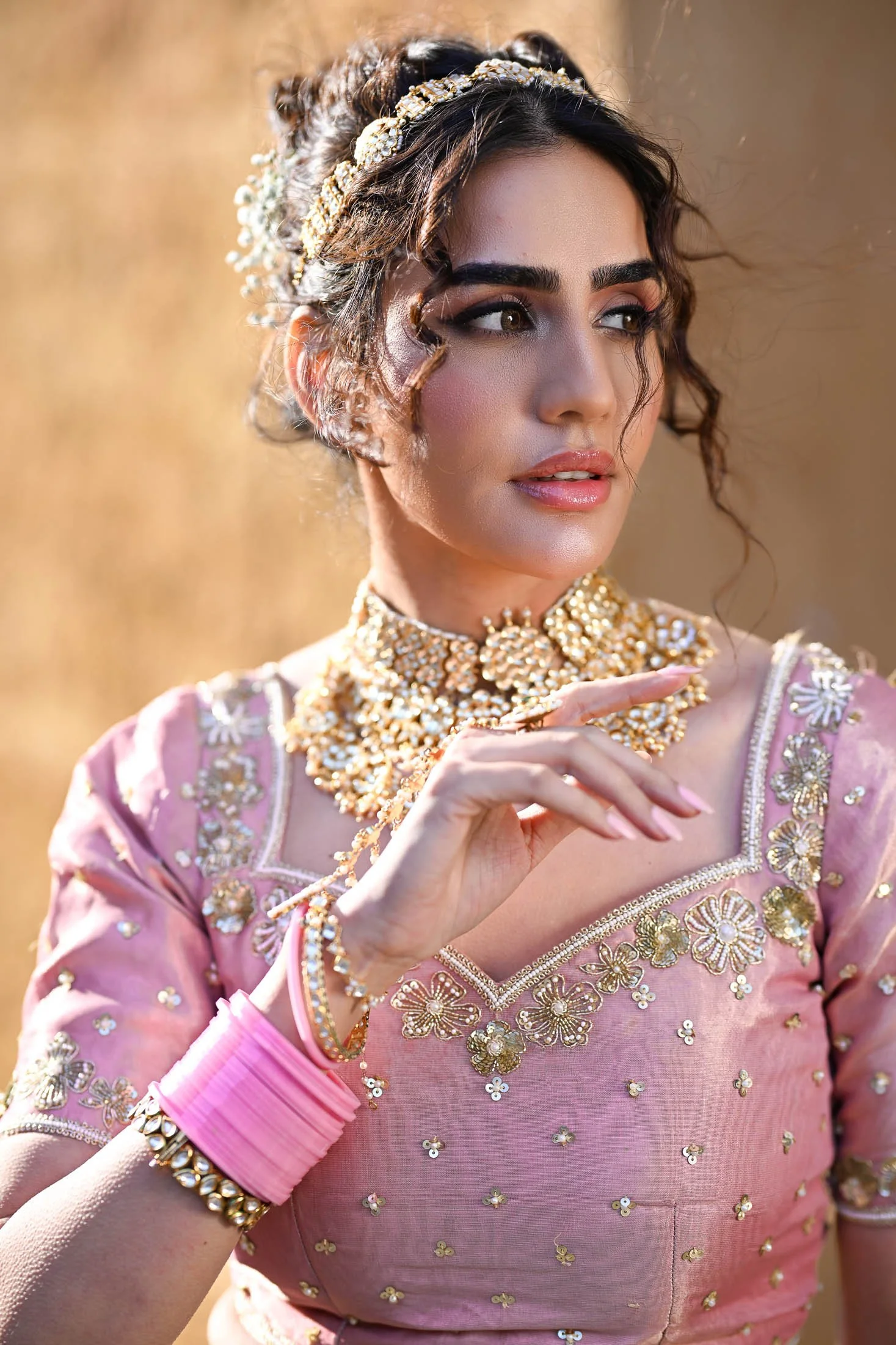 A woman dressed in traditional Indian attire, wearing intricate gold jewelry including a choker necklace, earrings, and bangles, with a pink embroidered dress. She has styled hair with a headband and is posing outdoors with a blurred background and w