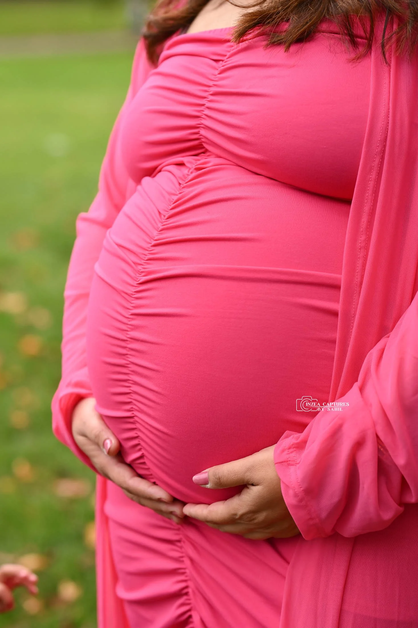 Close-up of a pregnant woman in a pink dress holding her belly with both hands, outdoors in a grassy area.