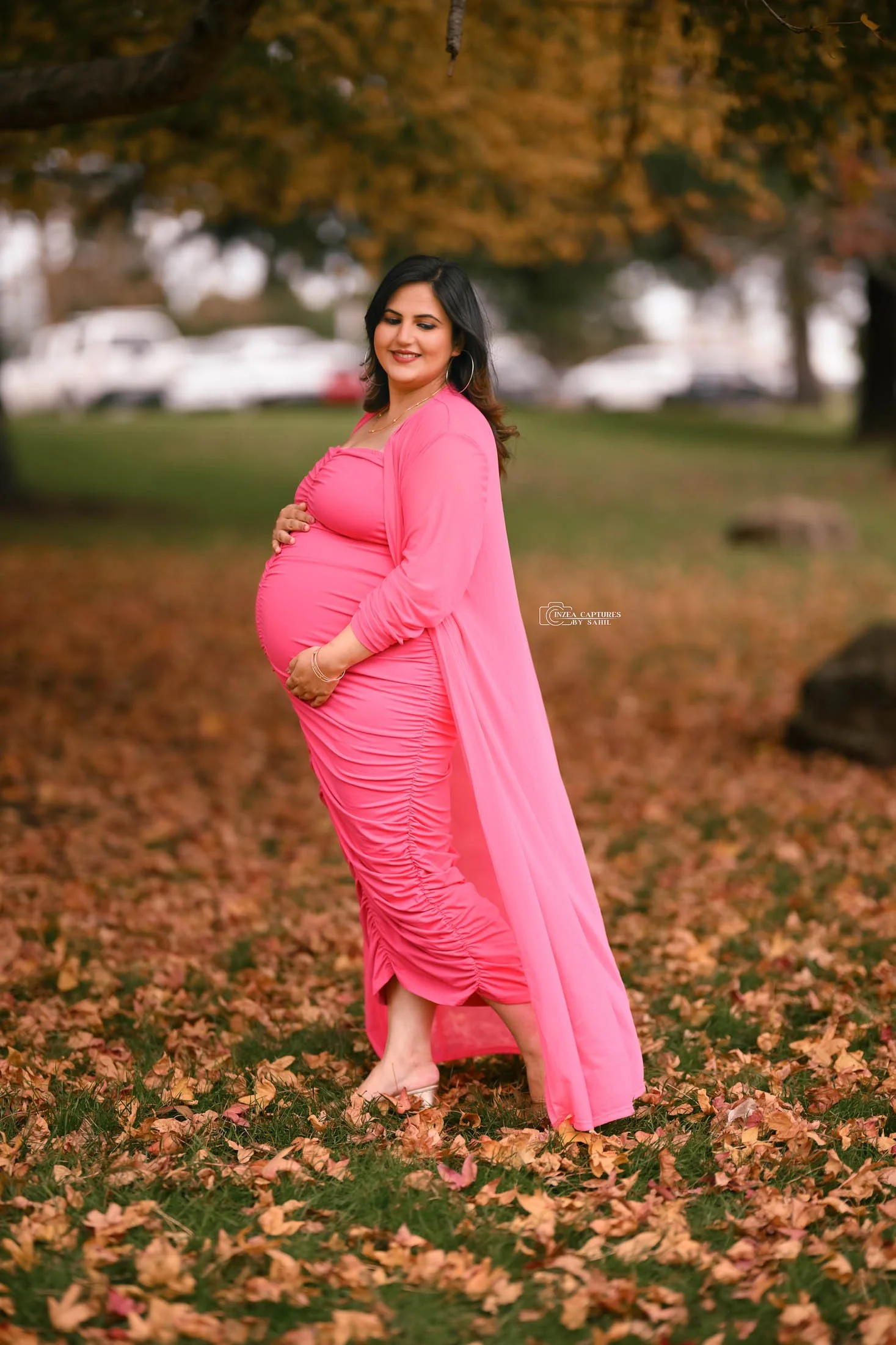Pregnant woman in a bright pink dress and matching cover-up standing barefoot on fallen autumn leaves in a park, smiling with hand on stomach and looking down.
