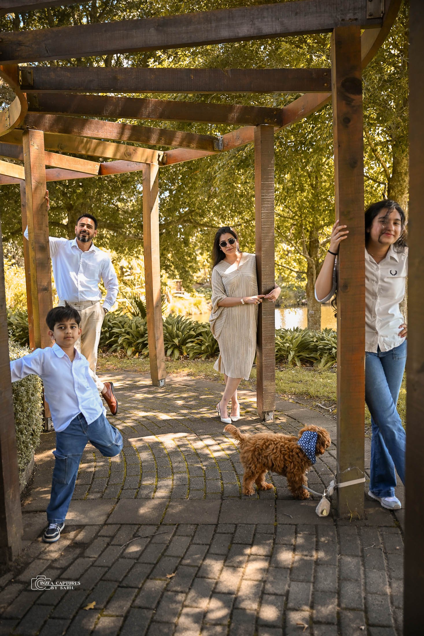 A family of five with a dog standing under a wooden pergola in a park on a sunny day. The father, mother, and three children are posing, with the dog sitting on the ground.