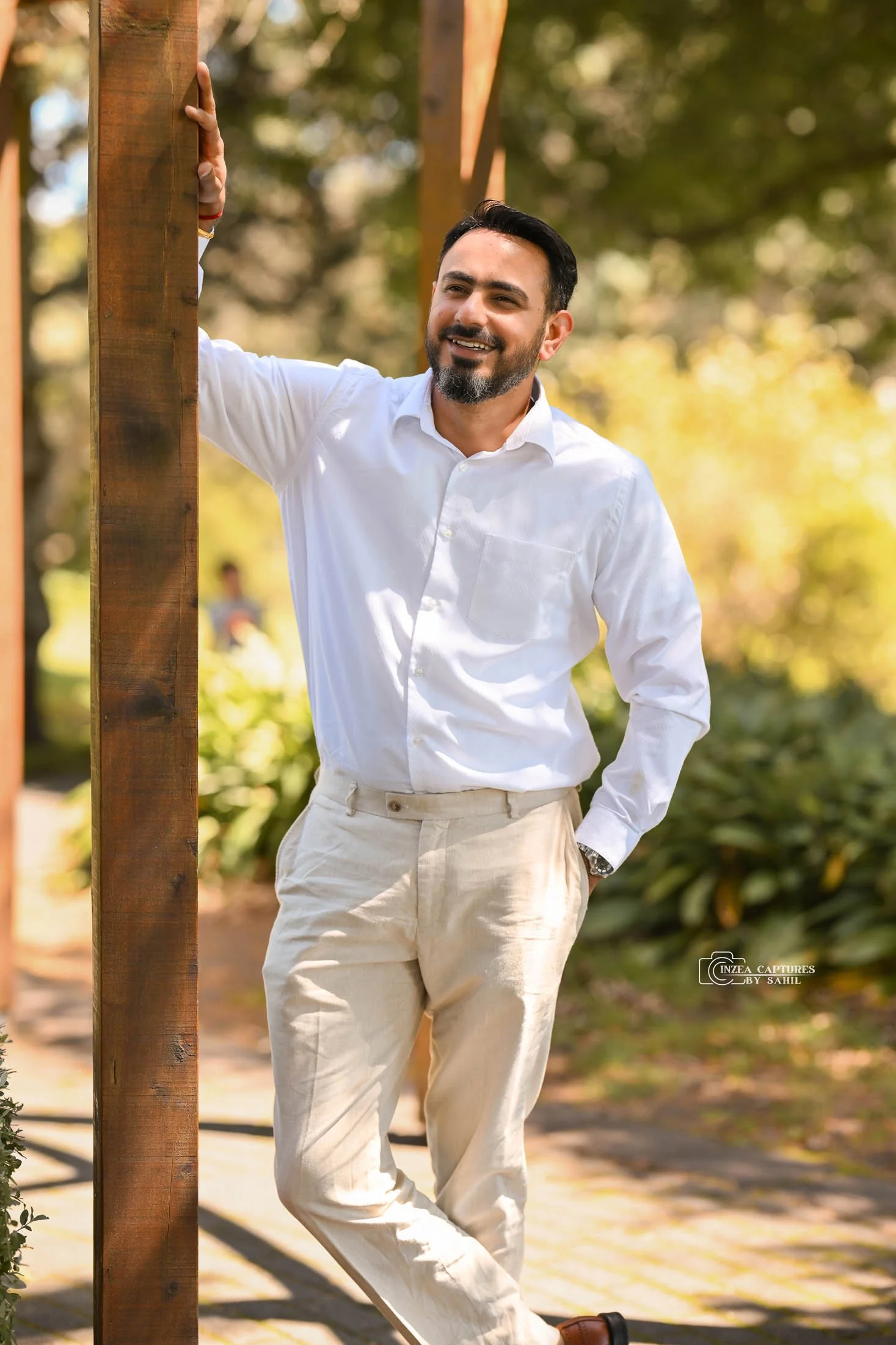 A man with short dark hair and a beard, wearing a white shirt and beige pants, smiling and standing outdoors in a park during daytime, leaning against a wooden post.