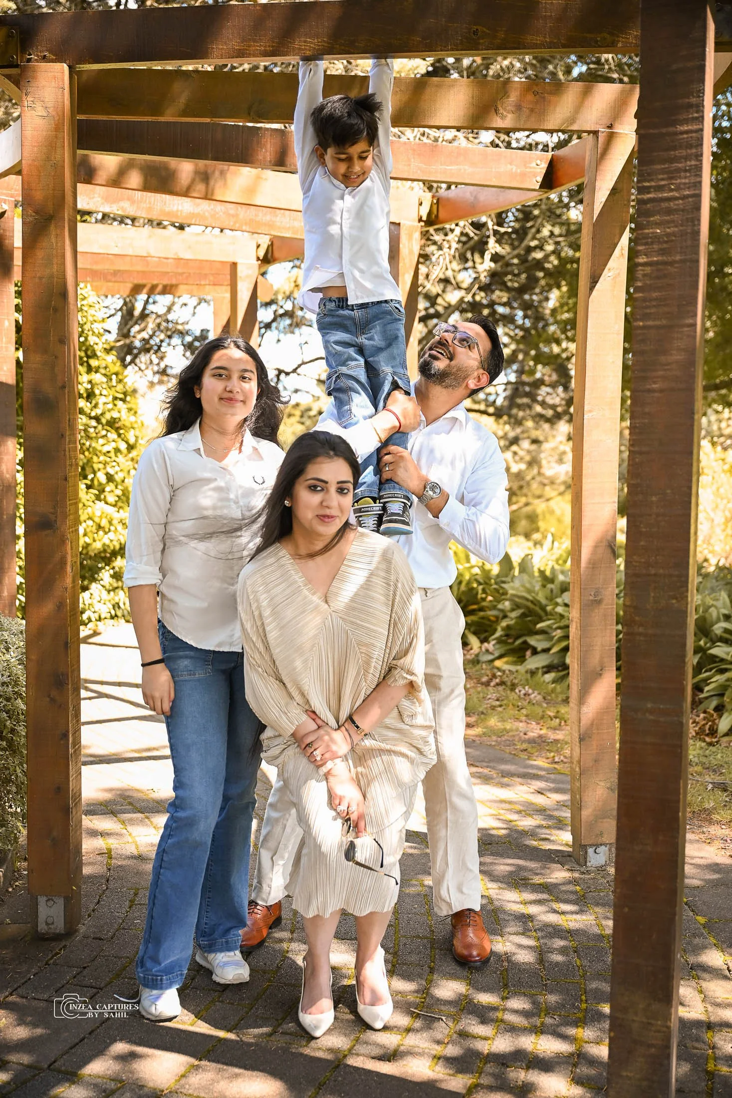 A group of five people outdoors, with one man helping a young boy hang from a wooden structure while the others pose nearby. The setting is a sunny park or garden with trees and greenery.