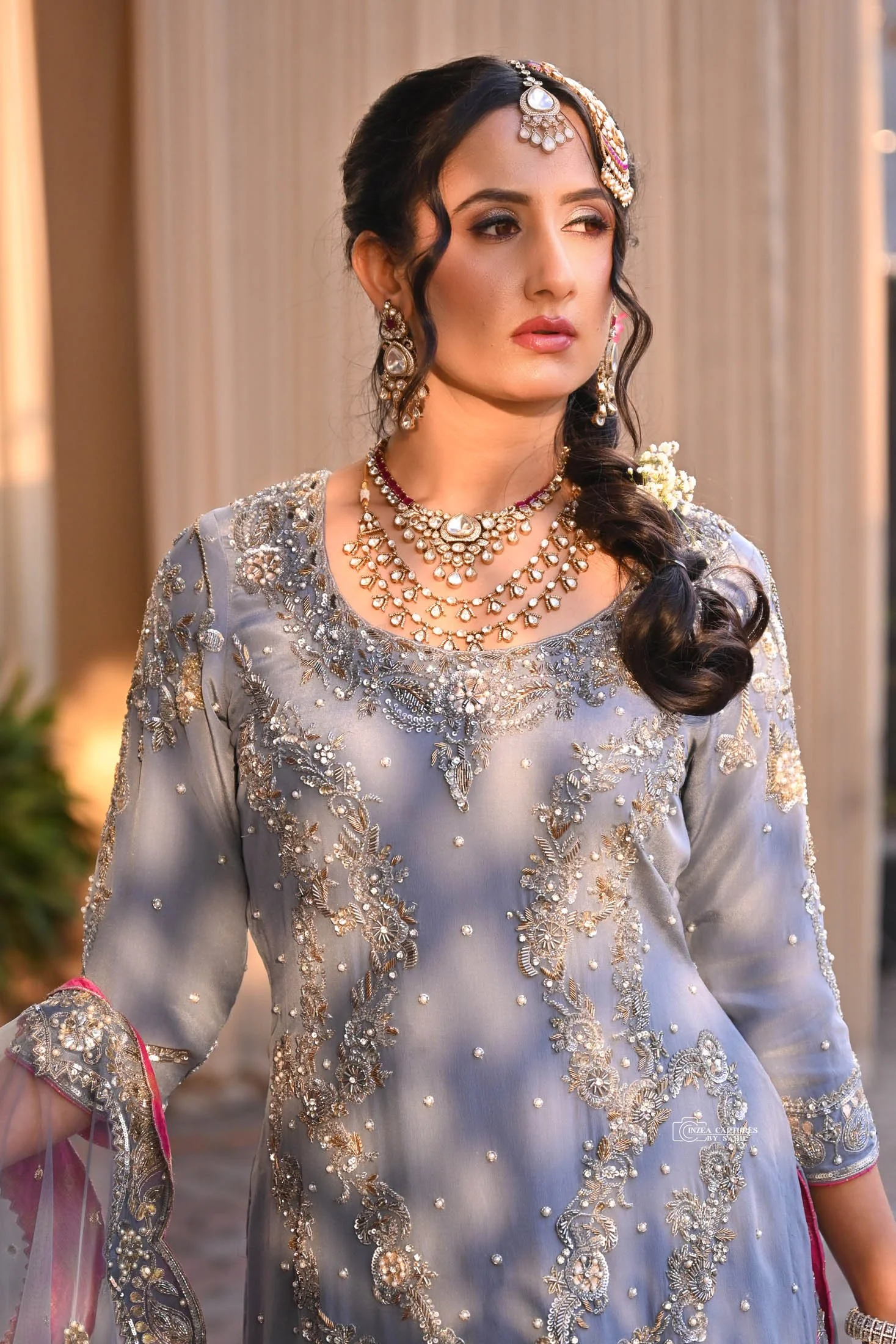 A woman wearing a glamorous gray traditional dress with intricate silver embroidery and jewelry, set indoors with columns in the background.