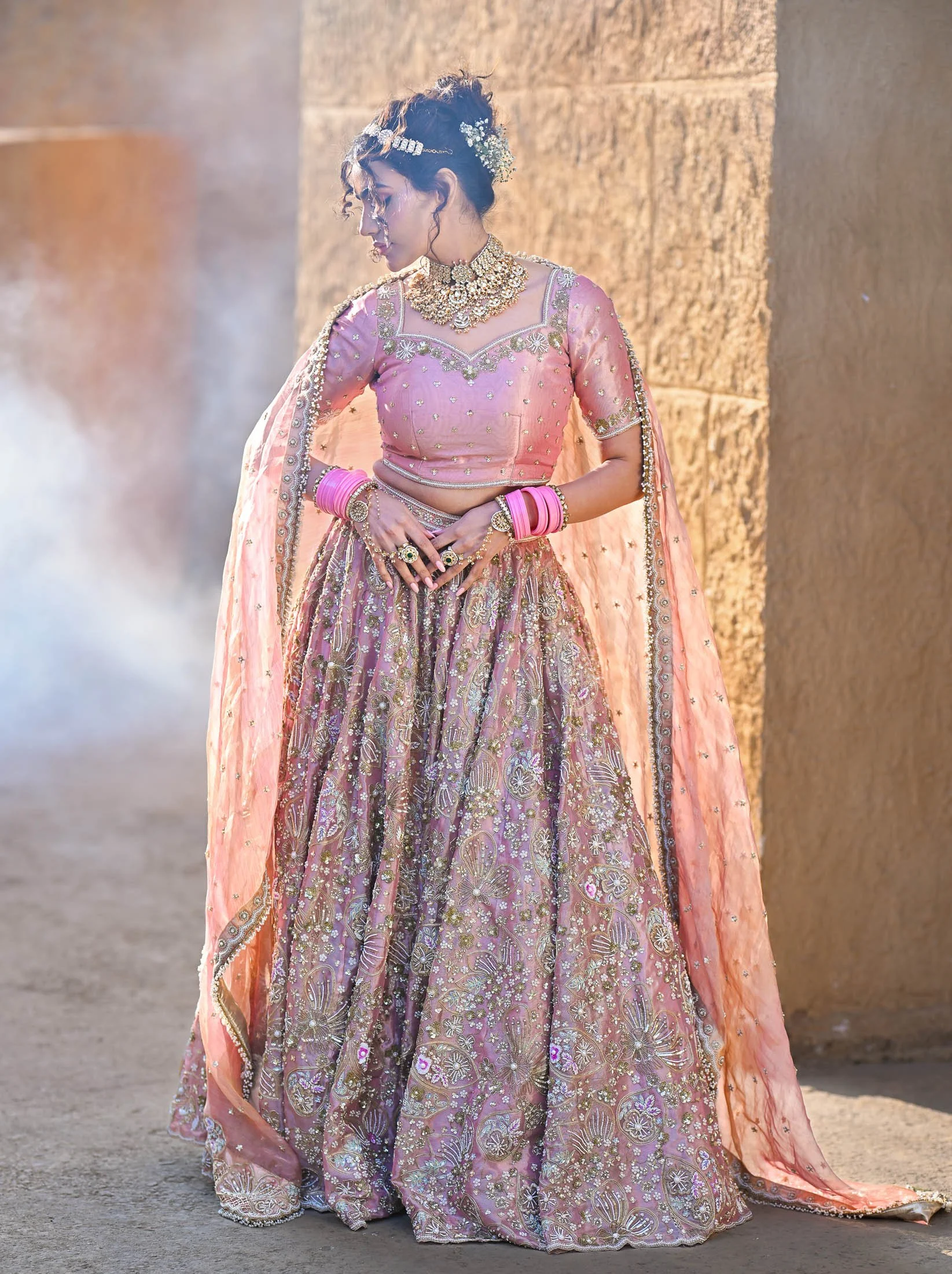 A woman dressed in a traditional pink and gold embroidered lehenga with matching dupatta, adorned with jewelry and bangles, standing against a stone wall with smoke and a soft light in the background.