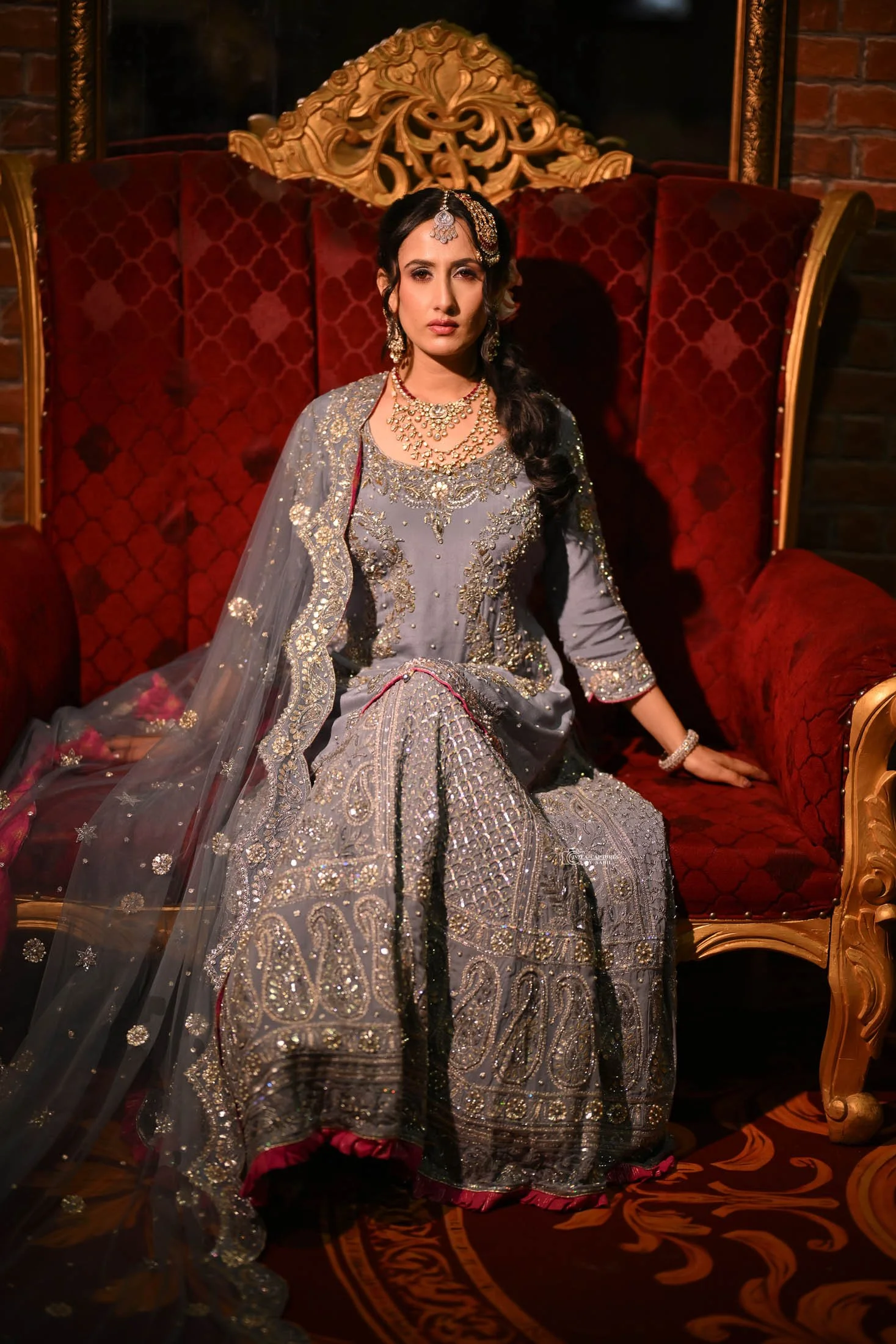 A woman dressed in traditional South Asian bridal attire, seated on a luxurious red and gold ornate sofa, surrounded by a dark, elegant interior.