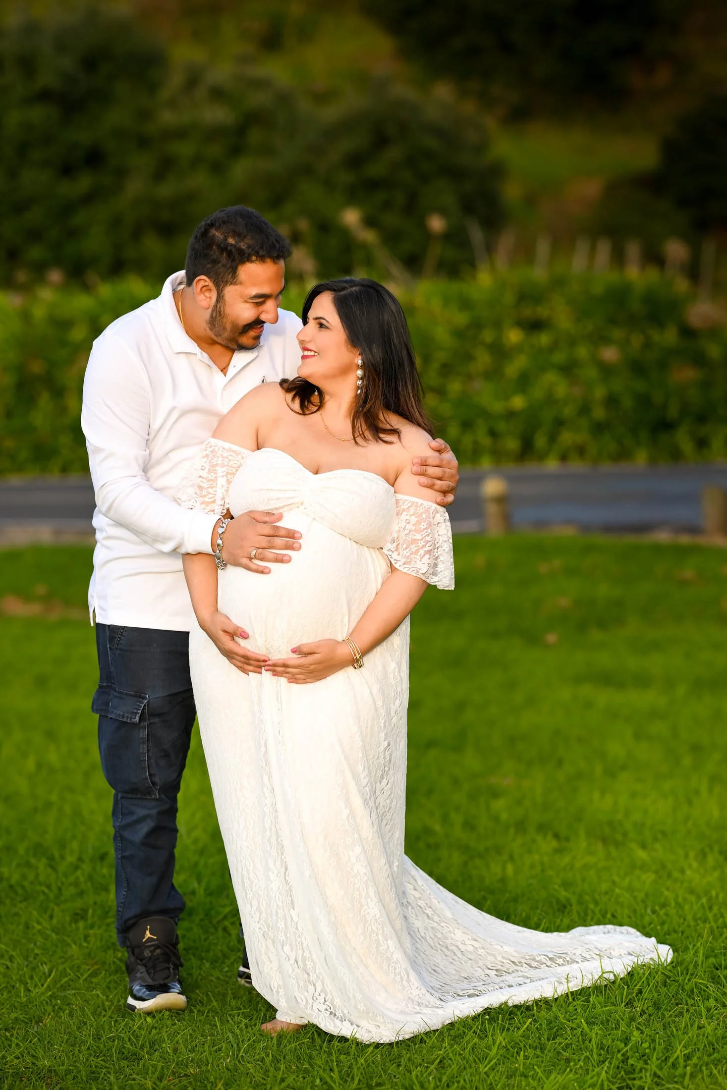 A pregnant woman in a white lace dress standing on grass, with a man in a white shirt and dark jeans embracing her from behind in an outdoor setting with green trees in the background.