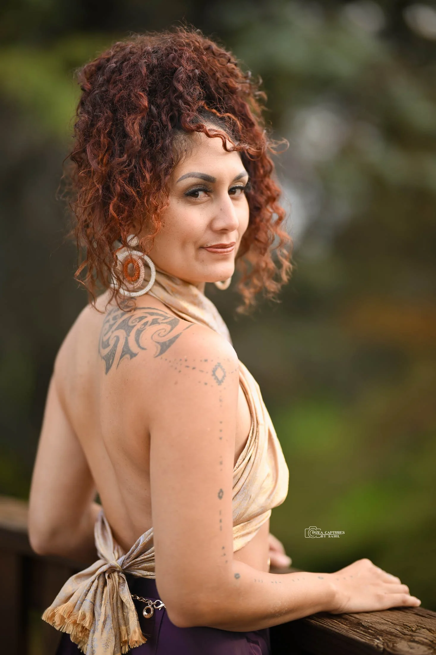 A woman with curly red hair, tattoos on her shoulder and arm, wearing large earrings and a sleeveless top, standing outdoors near a wooden railing, looking at the camera against a blurred green background.