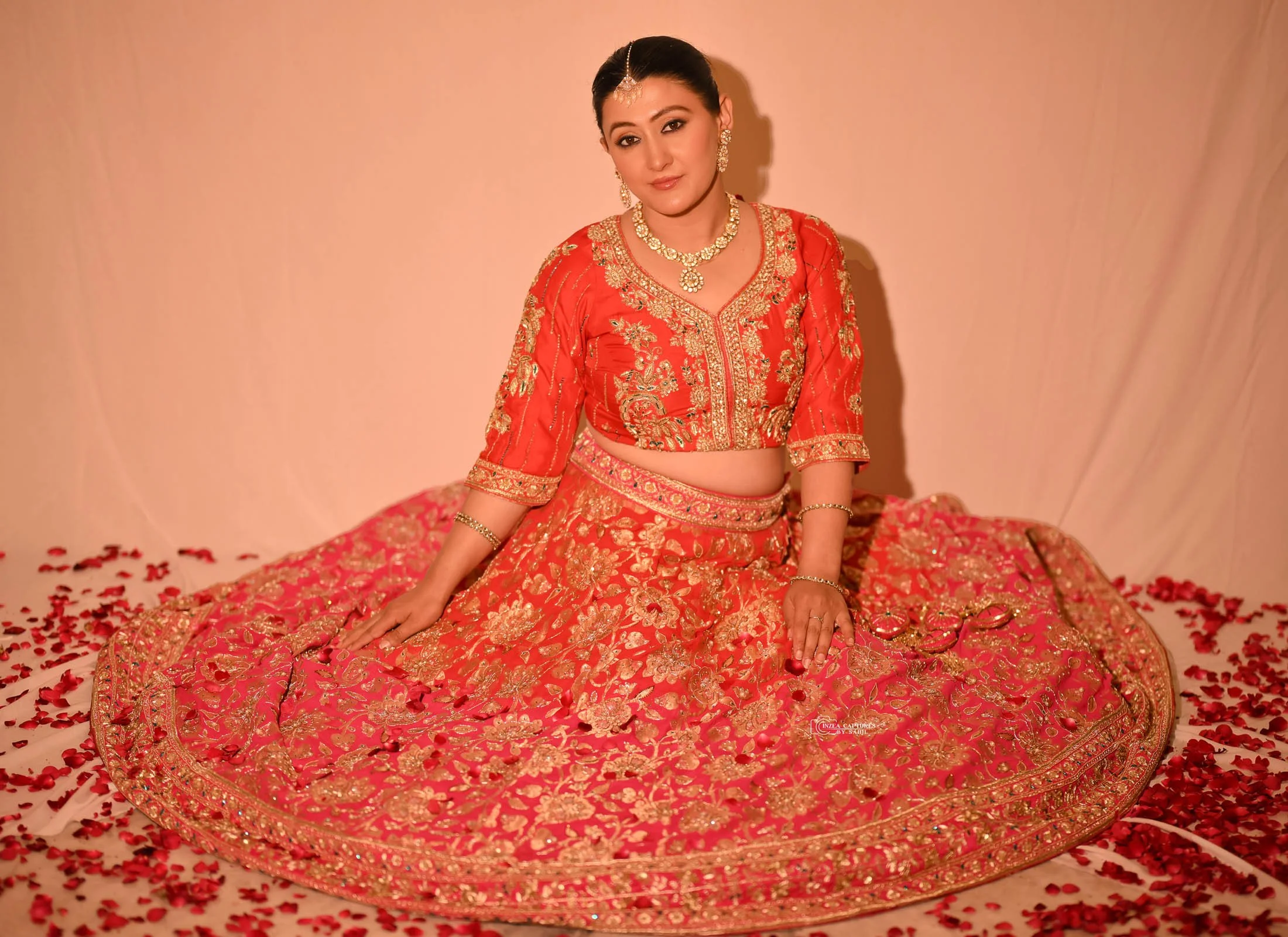 A woman dressed in a red and gold traditional Indian outfit, sitting on the floor surrounded by rose petals with jewelry including earrings, necklace, bracelet, and a headpiece.