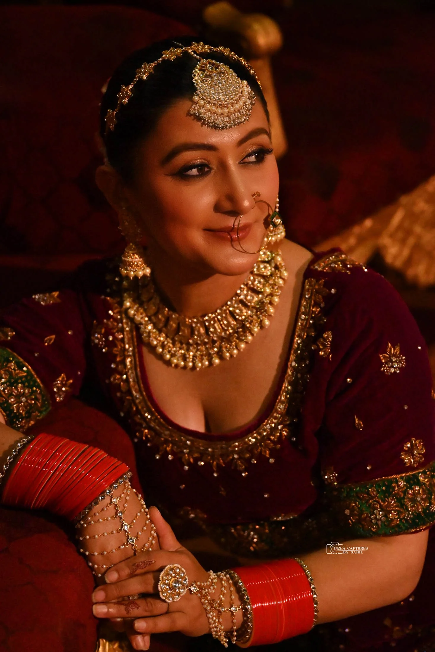 Woman in traditional Indian attire wearing jewelry, including necklace, earrings, nose ring, headpiece, and bangles, sitting with her arms crossed on a red armrest.