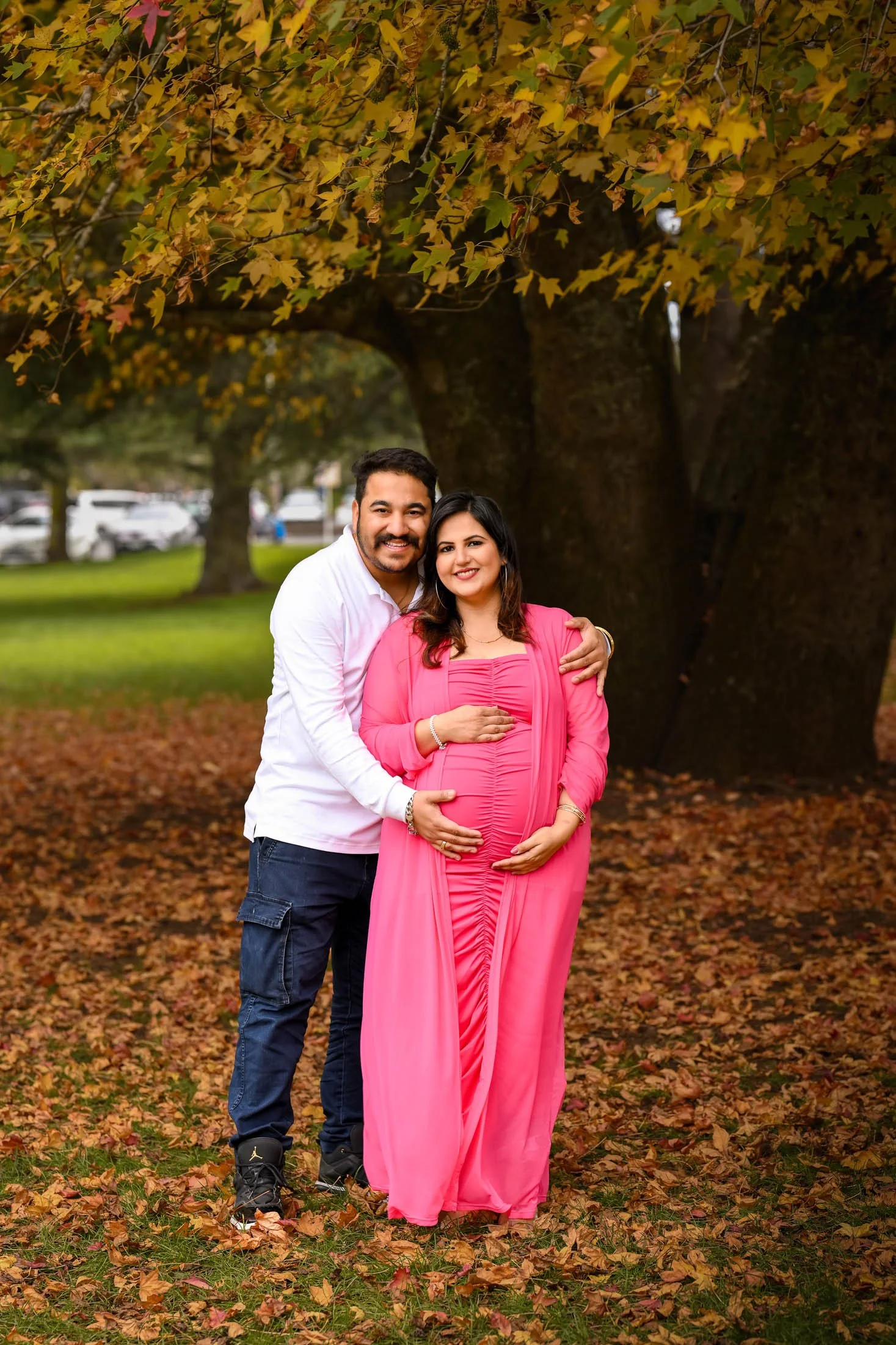 A pregnant woman in a bright pink dress and a man in a white shirt stand together in a park during autumn, with large trees and fallen leaves around them.