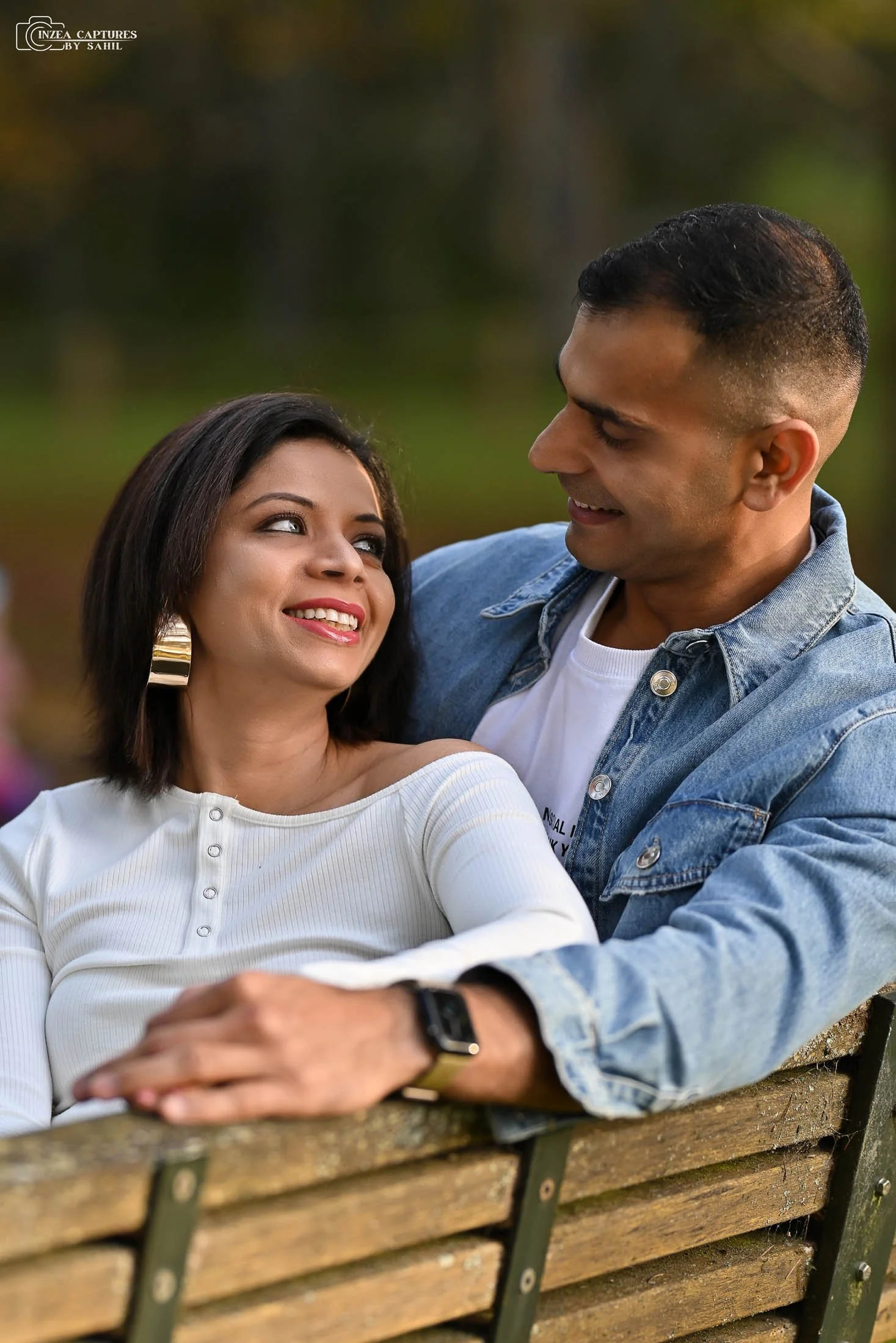 A man and woman sitting close together on a wooden park bench, smiling and looking at each other, with a blurred green background.