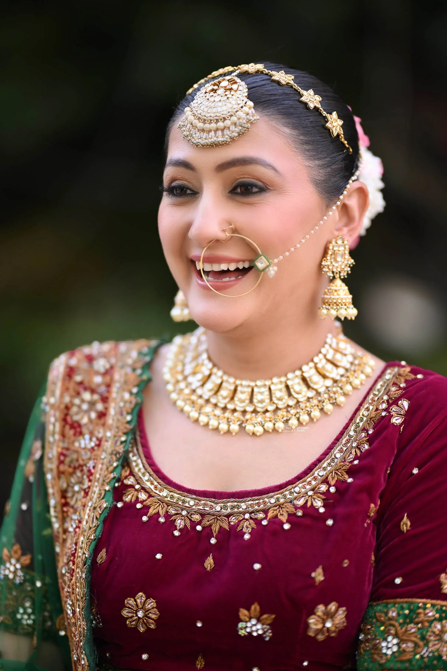 A woman dressed in traditional Indian bridal attire, smiling with gold jewelry and a velvet maroon and green outfit.