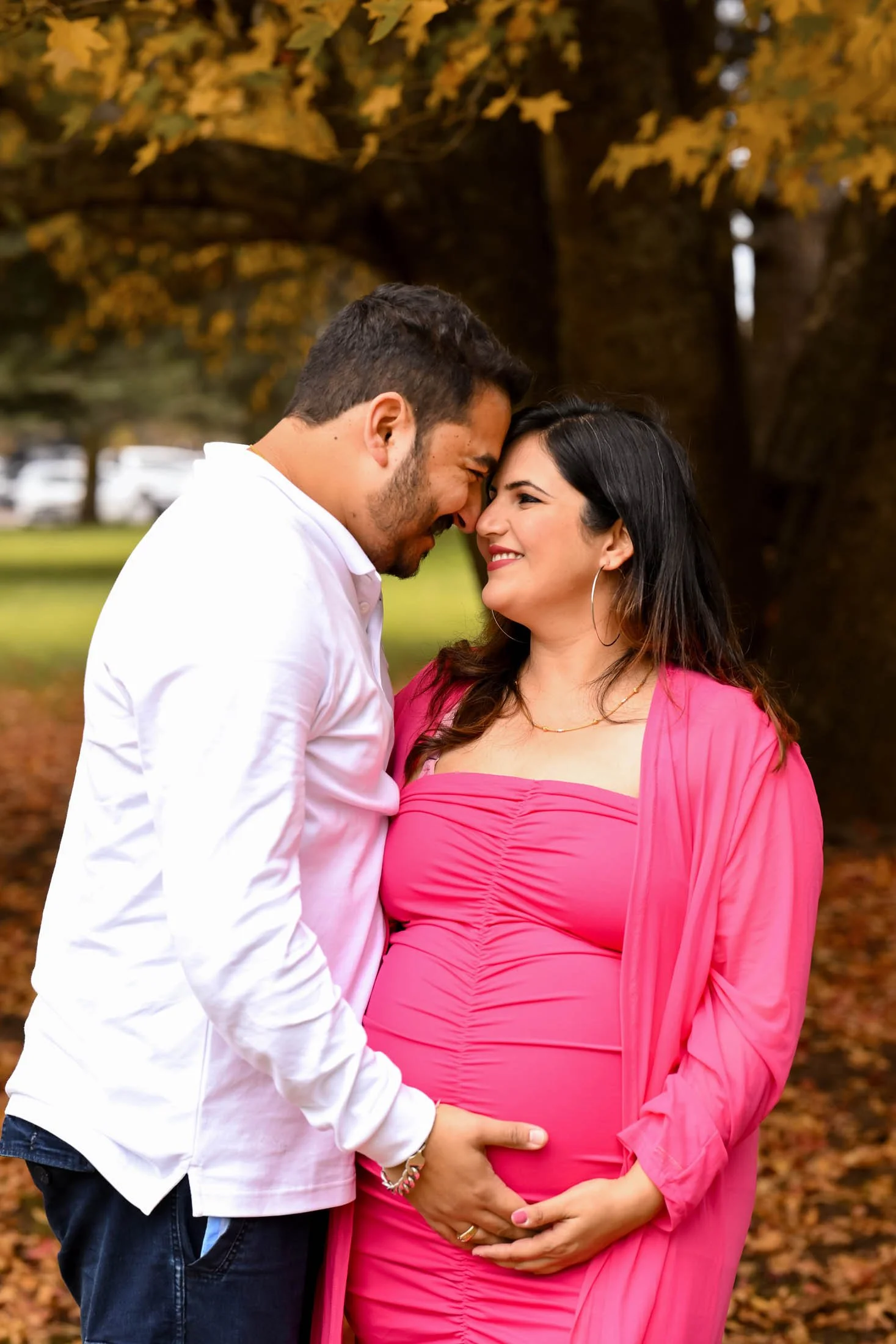 A couple, a man and a pregnant woman, standing close together outdoors, touching foreheads and holding her pregnant belly, with trees and autumn leaves in the background.