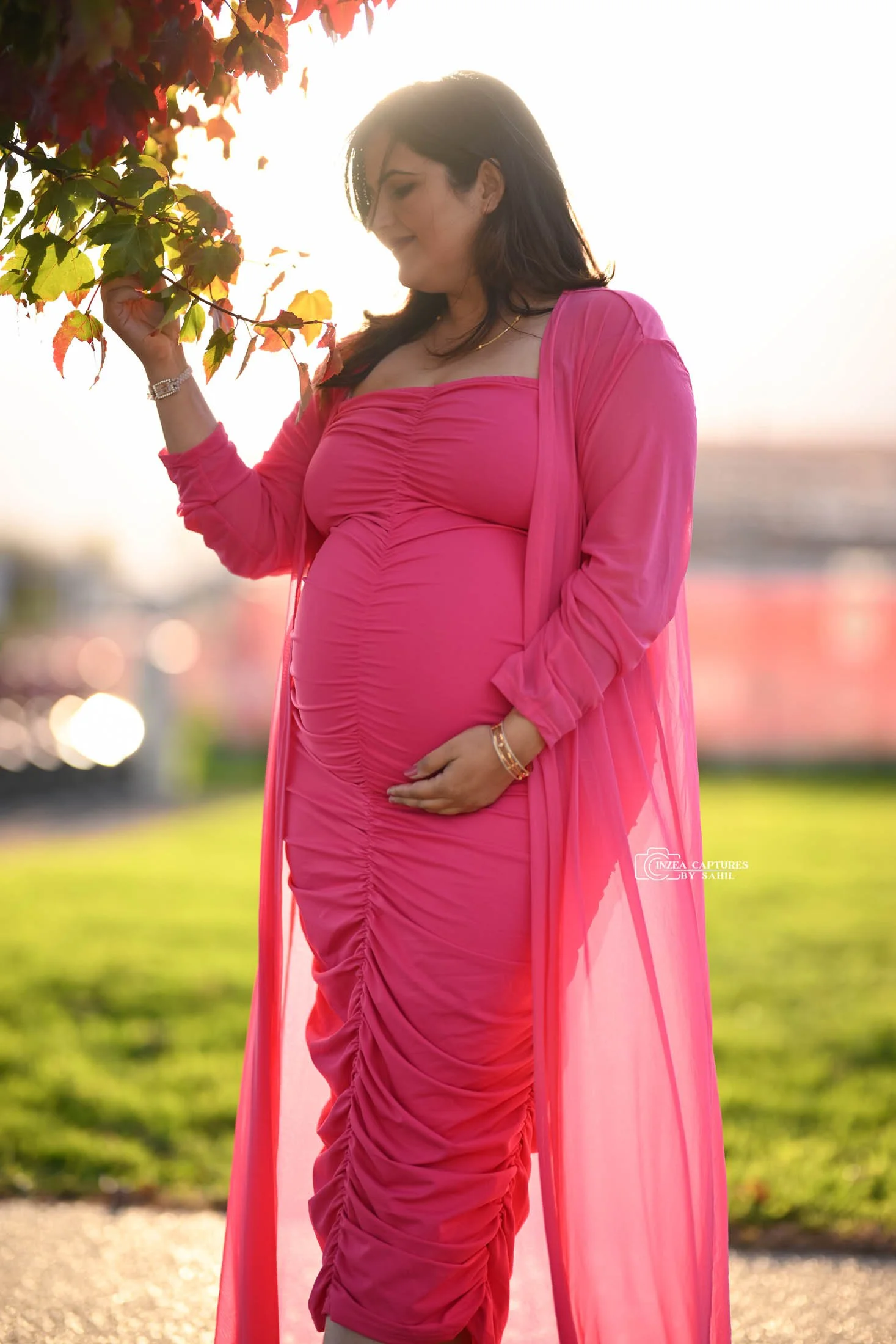 Pregnant woman in a bright pink dress and matching flowy cover-up standing outdoors near a tree, gently touching leaves with sunlight behind her.