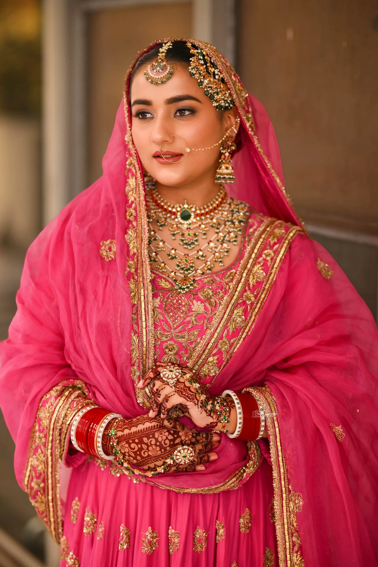 A woman dressed in traditional Indian bridal attire, wearing a pink saree with gold embroidery, ornate jewelry, and henna on her hands.