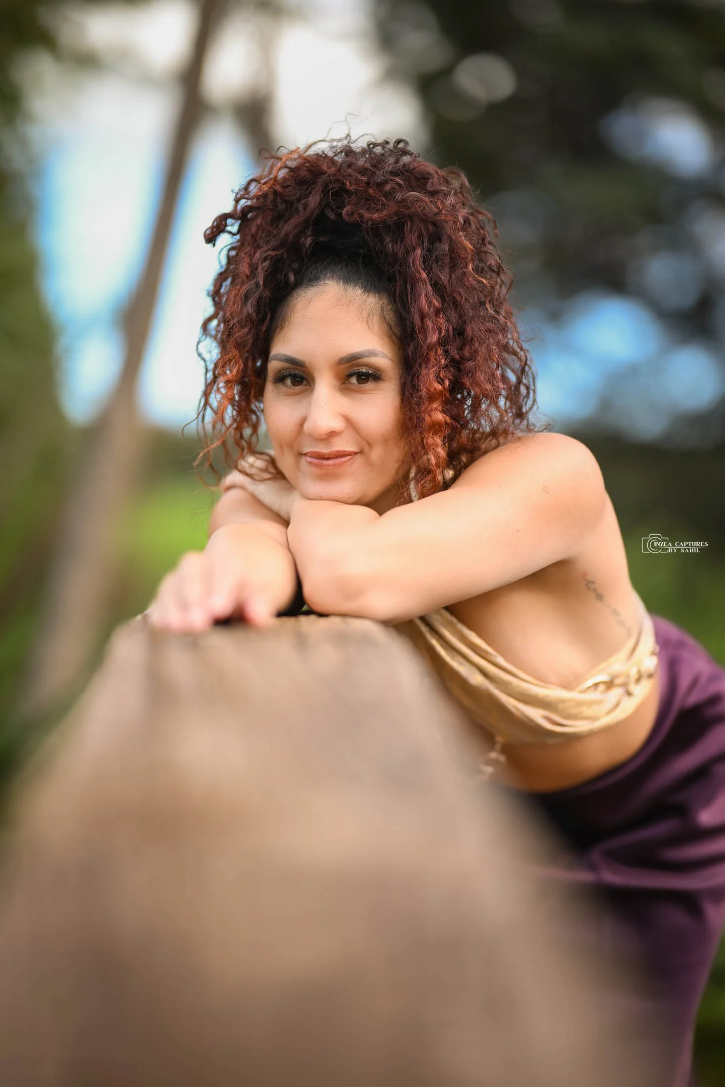 A woman with curly red hair leaning on a wooden surface outdoors, looking at the camera with a smile.