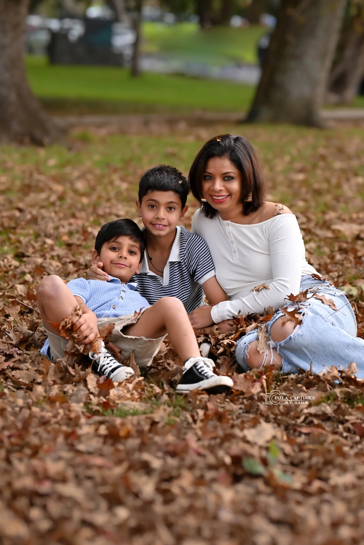A woman and two boys sitting on the ground covered in fallen leaves in a park, smiling at the camera.