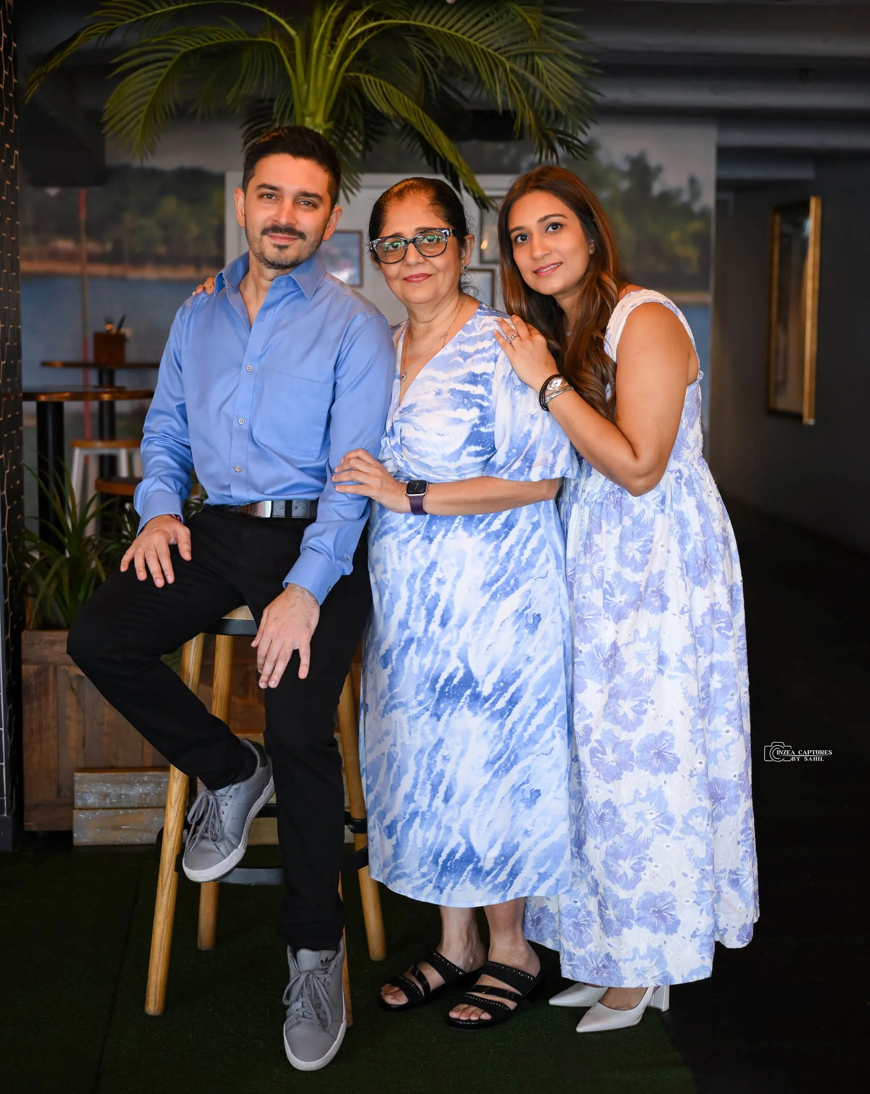 Three women and one man posing together indoors. The man is sitting on a stool, wearing a blue shirt and black pants. The woman in the middle is standing, wearing a blue and white patterned dress and glasses. The woman on the right is standing, weari