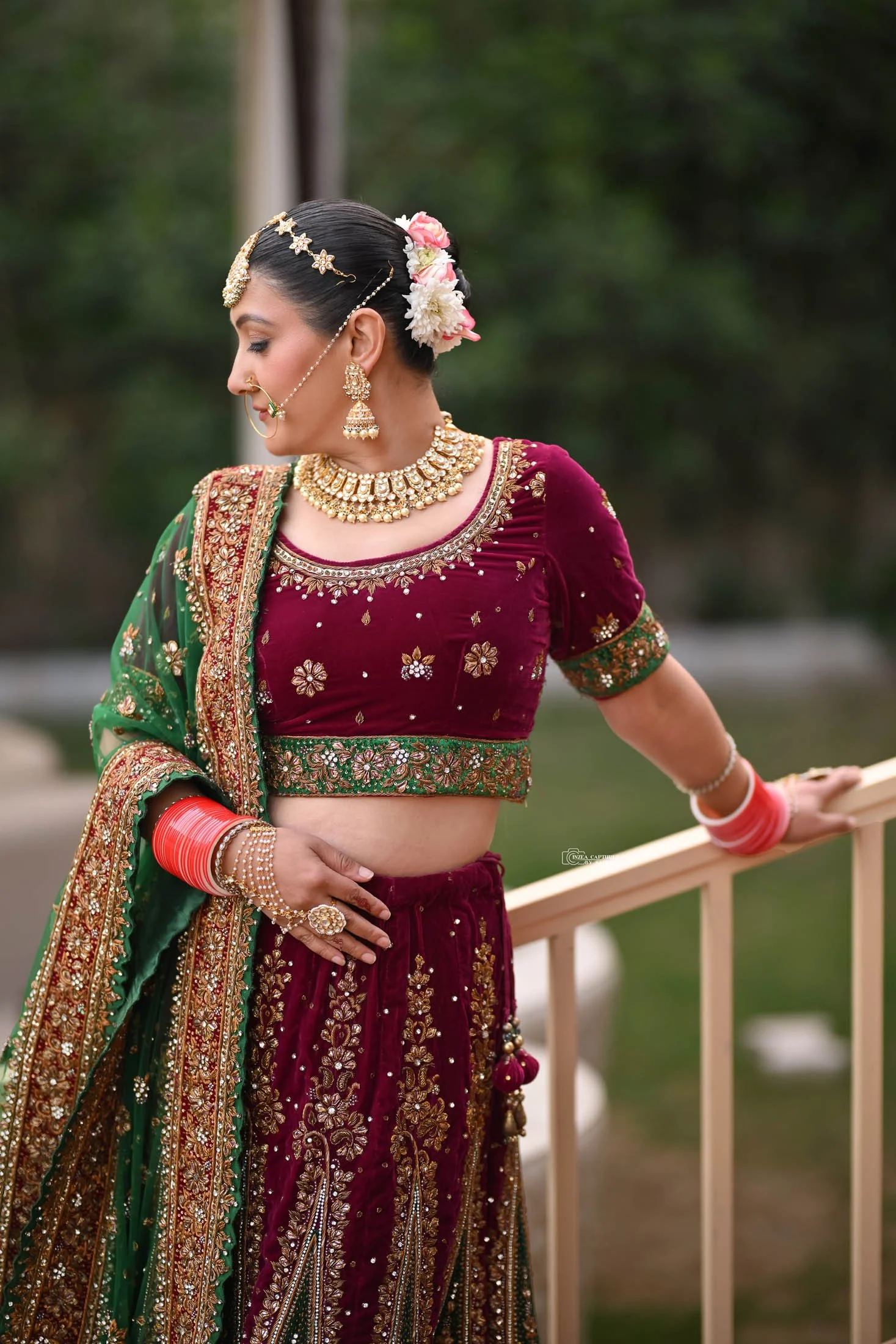 A woman wearing a traditional Indian maroon and green embroidered lehenga with gold jewelry, flowers in her hair, and red bangles, standing outdoors near a railing.