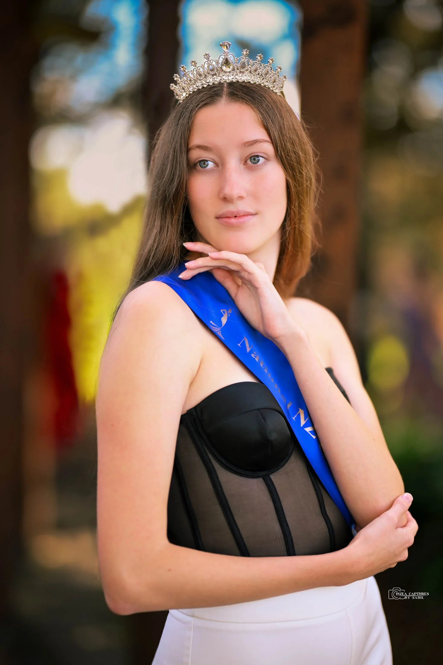 Young woman wearing a crown and a blue sash with a black strapless dress, posing outdoors with a blurred natural background.