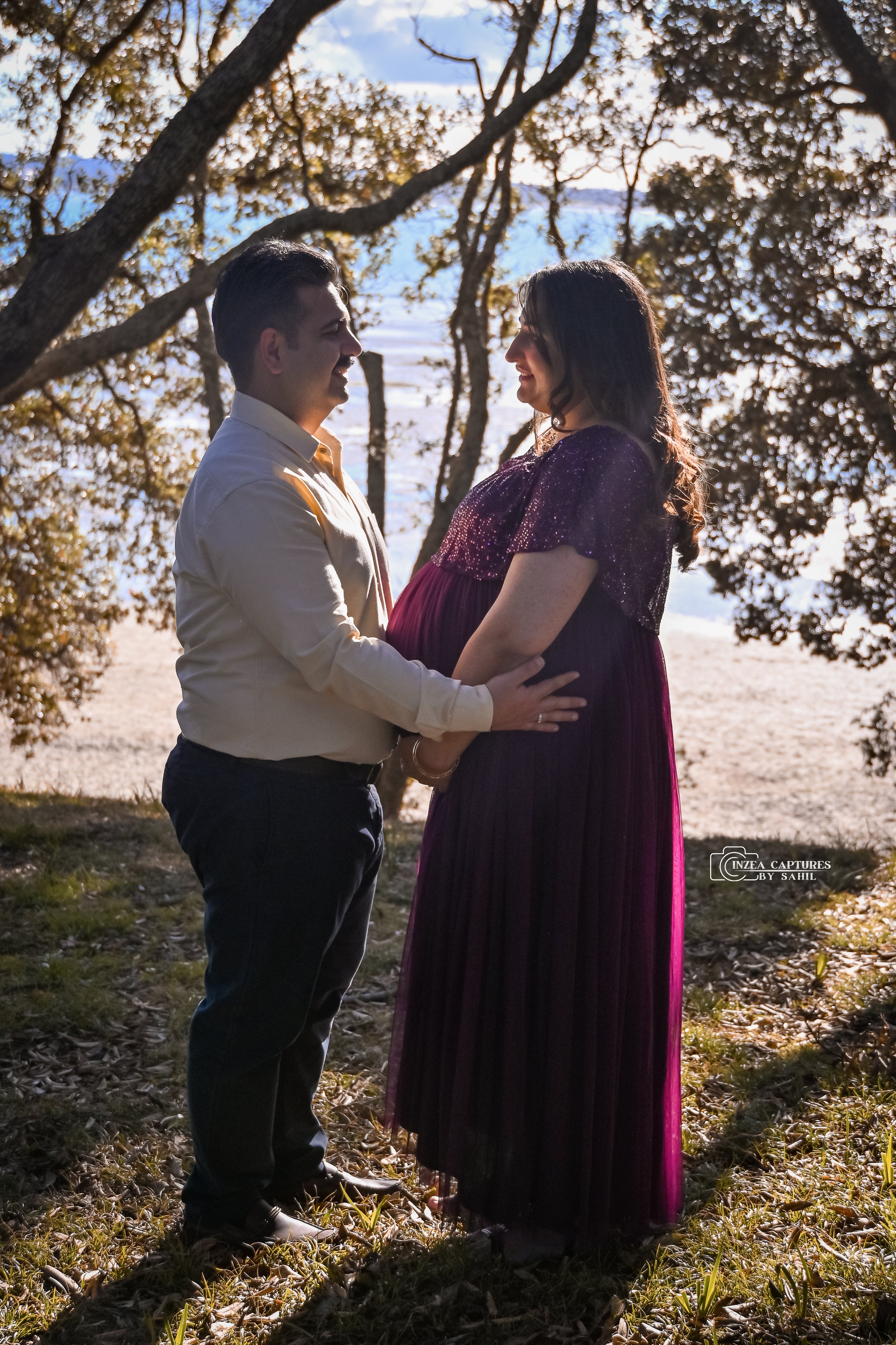 A couple, with the man holding a pregnant woman, standing outdoors under trees near a body of water during sunset, smiling at each other.