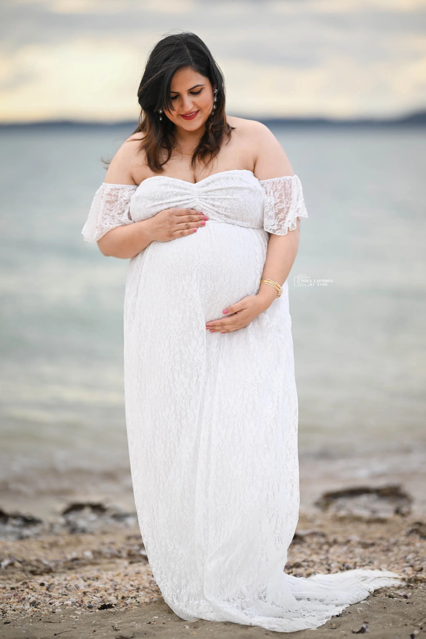 A pregnant woman in a white lace dress stands on a beach, smiling while gently touching her belly.