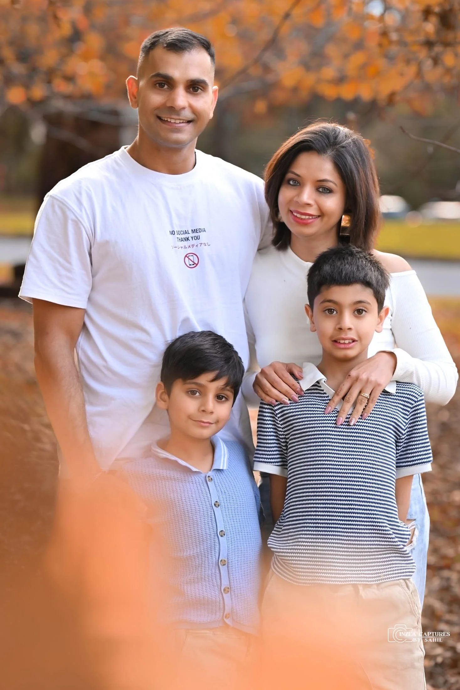 A family of four standing outdoors in an autumn setting with orange leaves in the background. The father is wearing a white t-shirt and smiling, the mother is wearing a white top, and the two sons are standing in front of them, one in a light purple 