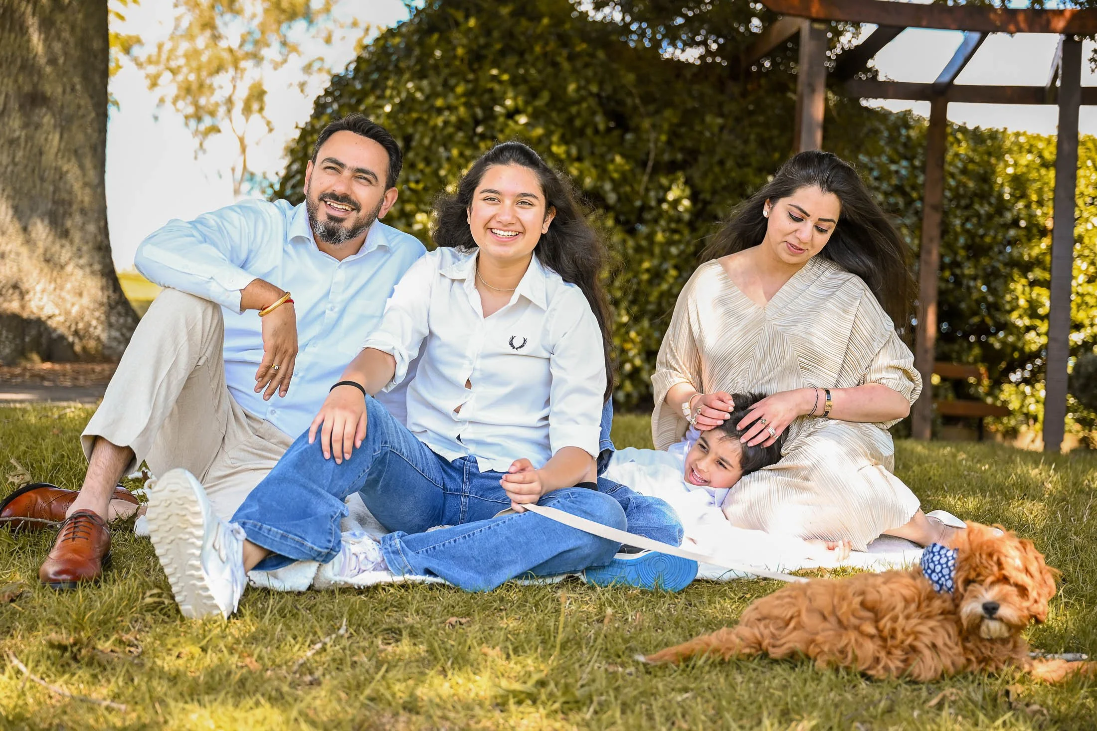 A family of four with a dog enjoying a sunny day outdoors on a grassy area, sitting and lying on a blanket.
