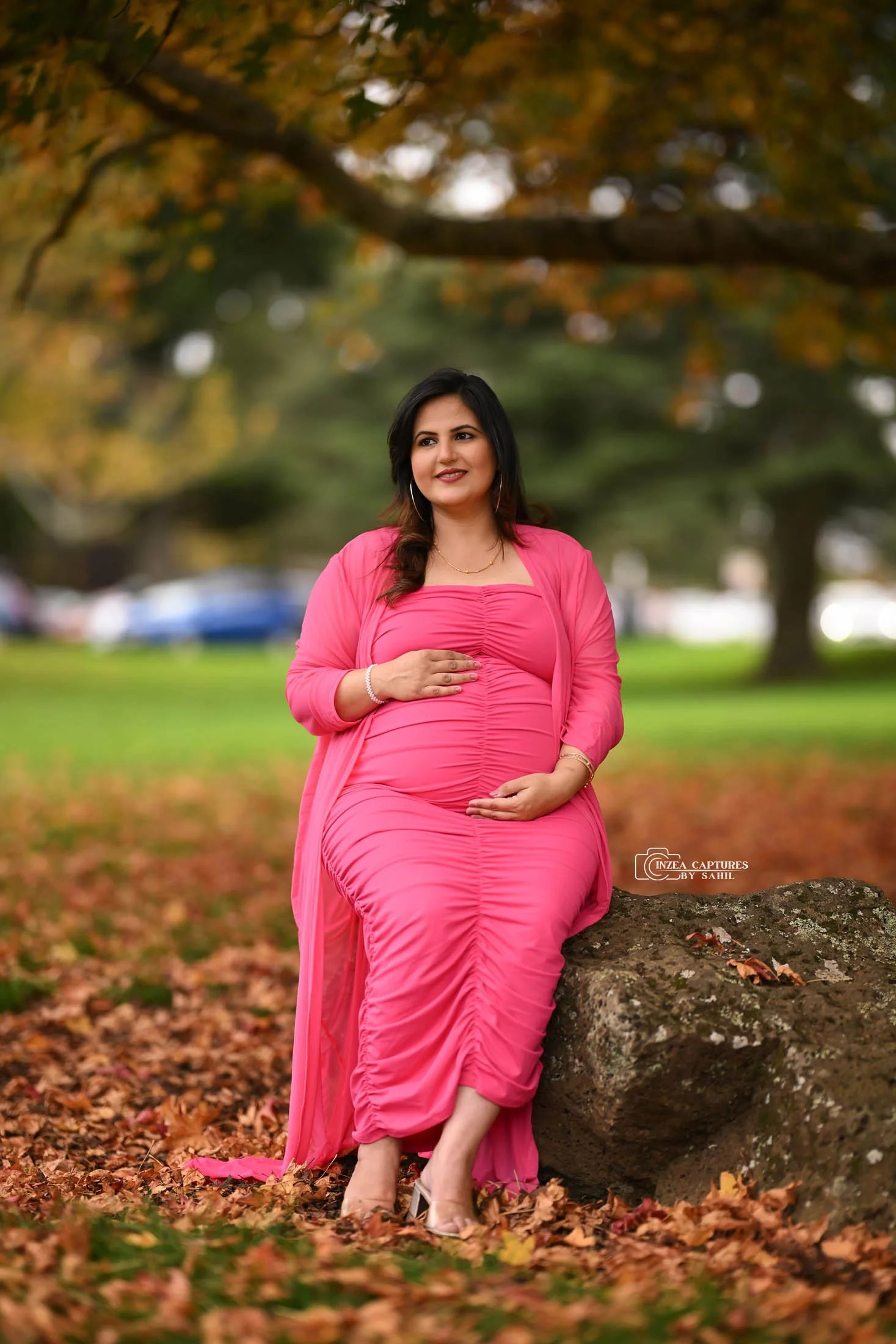 A pregnant woman in a bright pink dress and matching shawl sitting on a rock in a park, surrounded by fall leaves and trees.