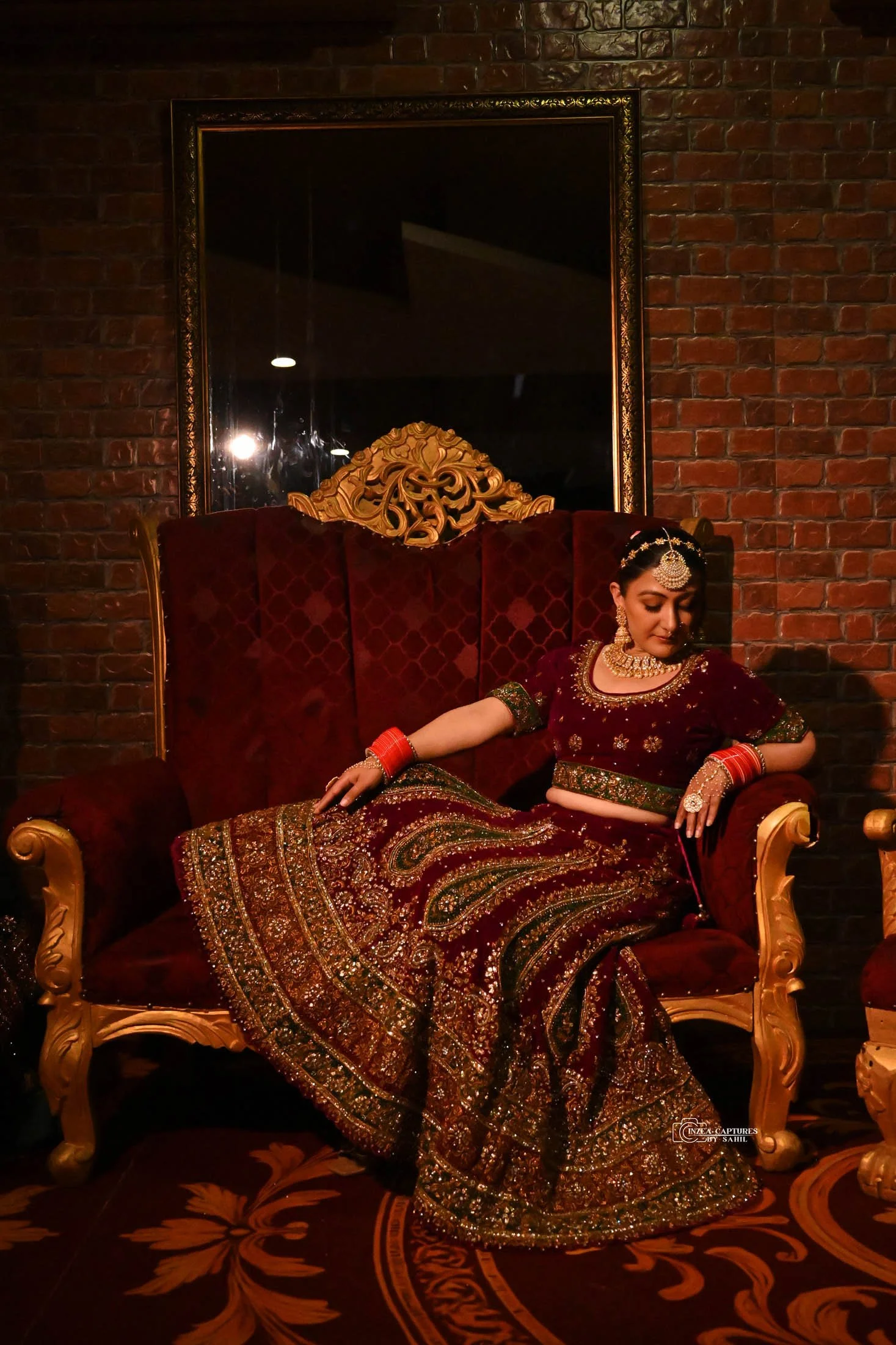 A woman dressed in traditional Indian attire, sitting on a red velvet sofa with ornate gold detailing, against a brick wall background.
