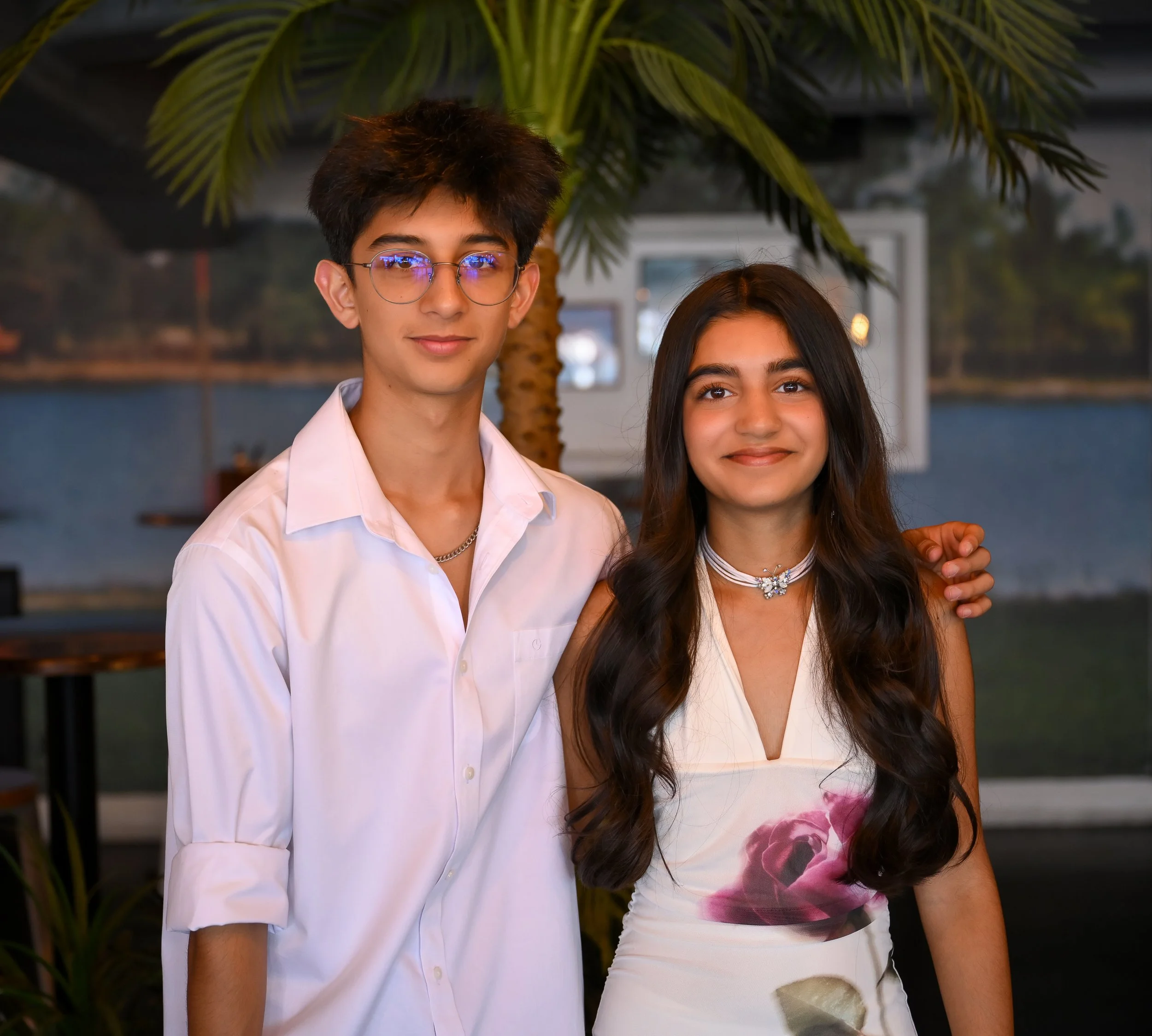 A young man and woman standing outdoors near a palm tree, smiling, with a body of water and a sky in the background.