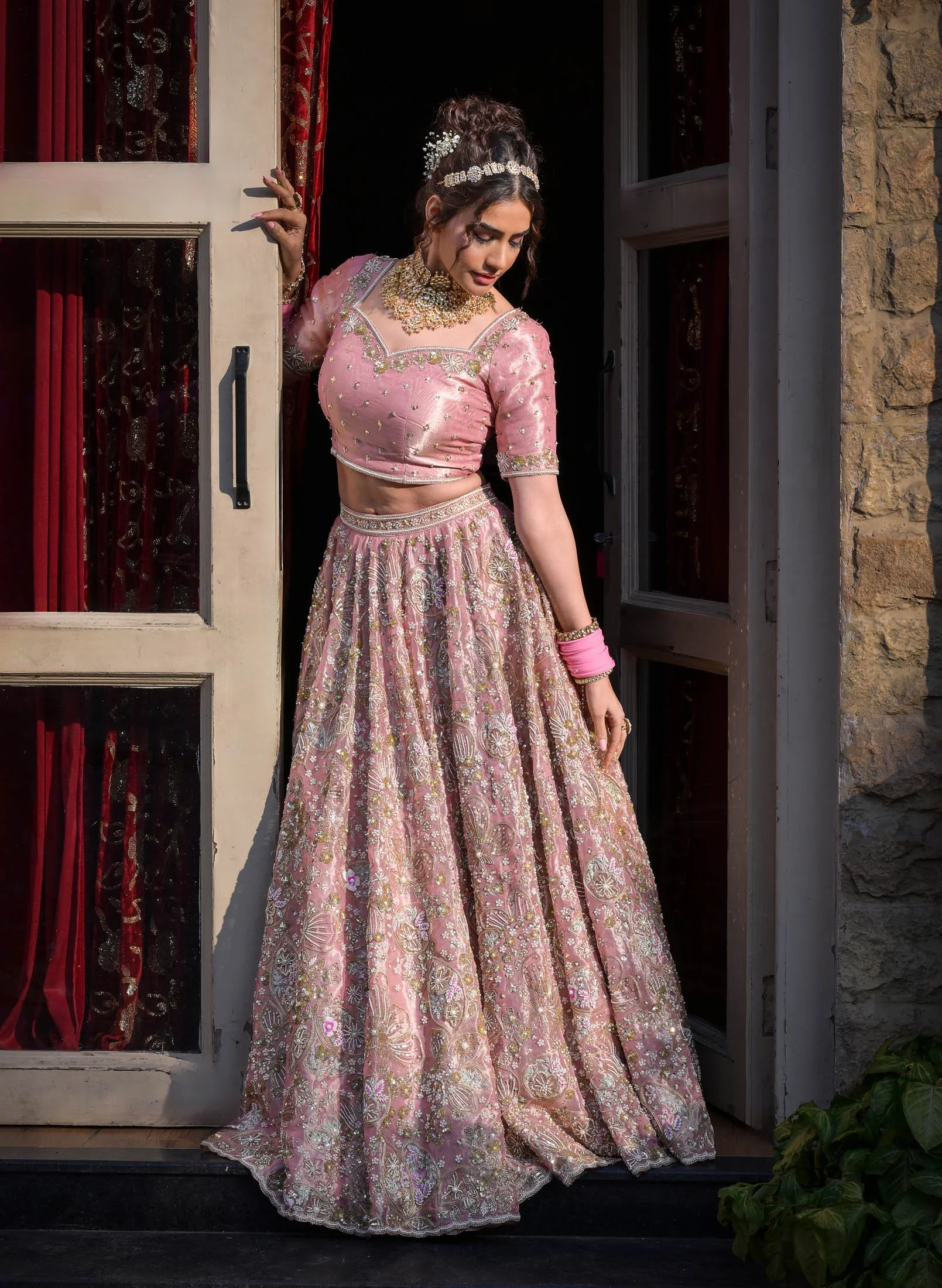 A young woman in traditional Indian attire, wearing a pink embroidered lehenga with gold and silver embellishments, poses near a doorway with red curtains. She has jewelry, including a necklace, earrings, and bangles, and her hair is styled with flow