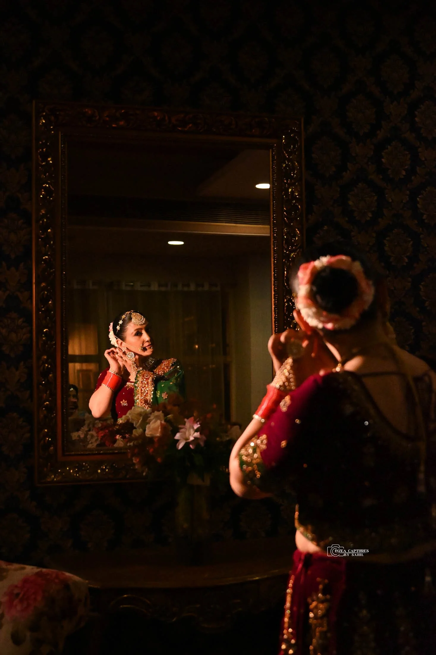 A woman in traditional Indian attire applies jewelry while looking in a mirror, with her reflection visible.