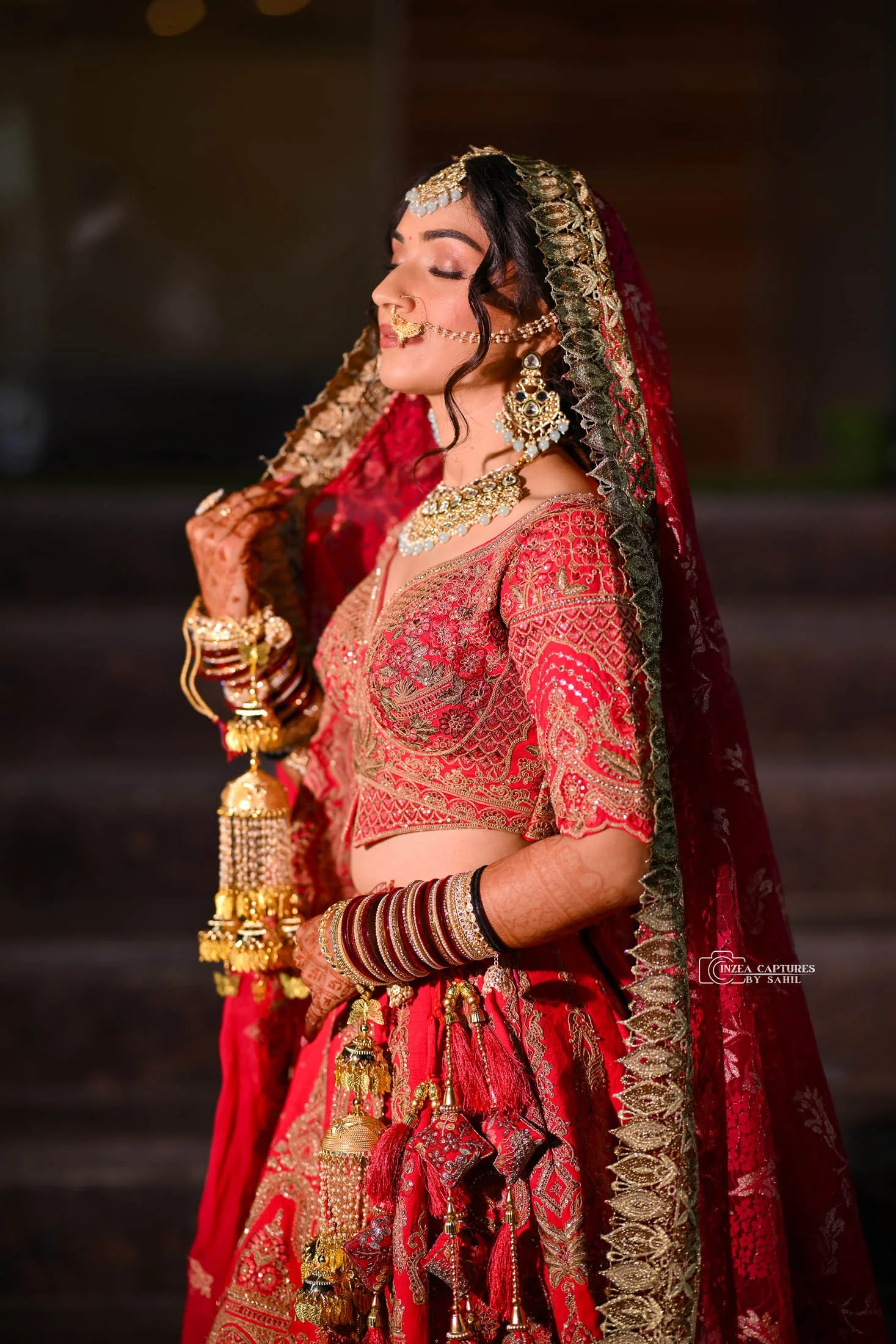 A woman in traditional Indian bridal attire, wearing a red and gold embroidered lehenga, with gold jewelry including a maang tikka, nose ring, earrings, necklace, and bangles, holding the edge of her veil with closed eyes.