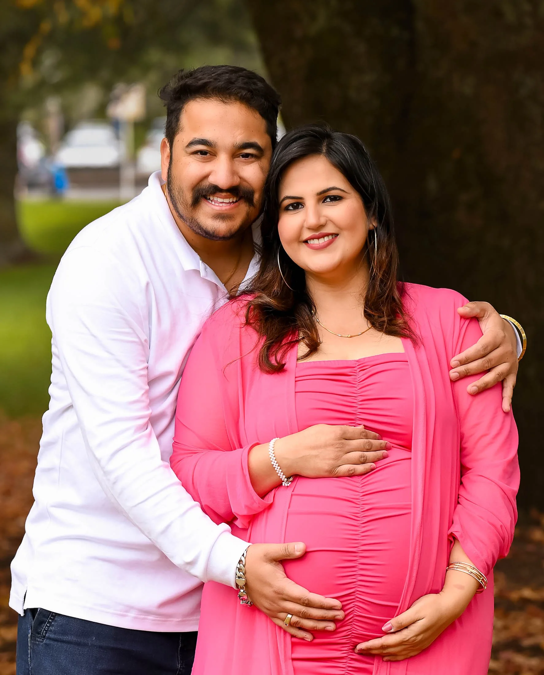 A smiling couple, a man and a pregnant woman, standing outside in a park during daytime, with trees in the background. The man is wearing a white shirt and has his arm around the woman's shoulder. The woman is wearing a pink dress and is cradling her
