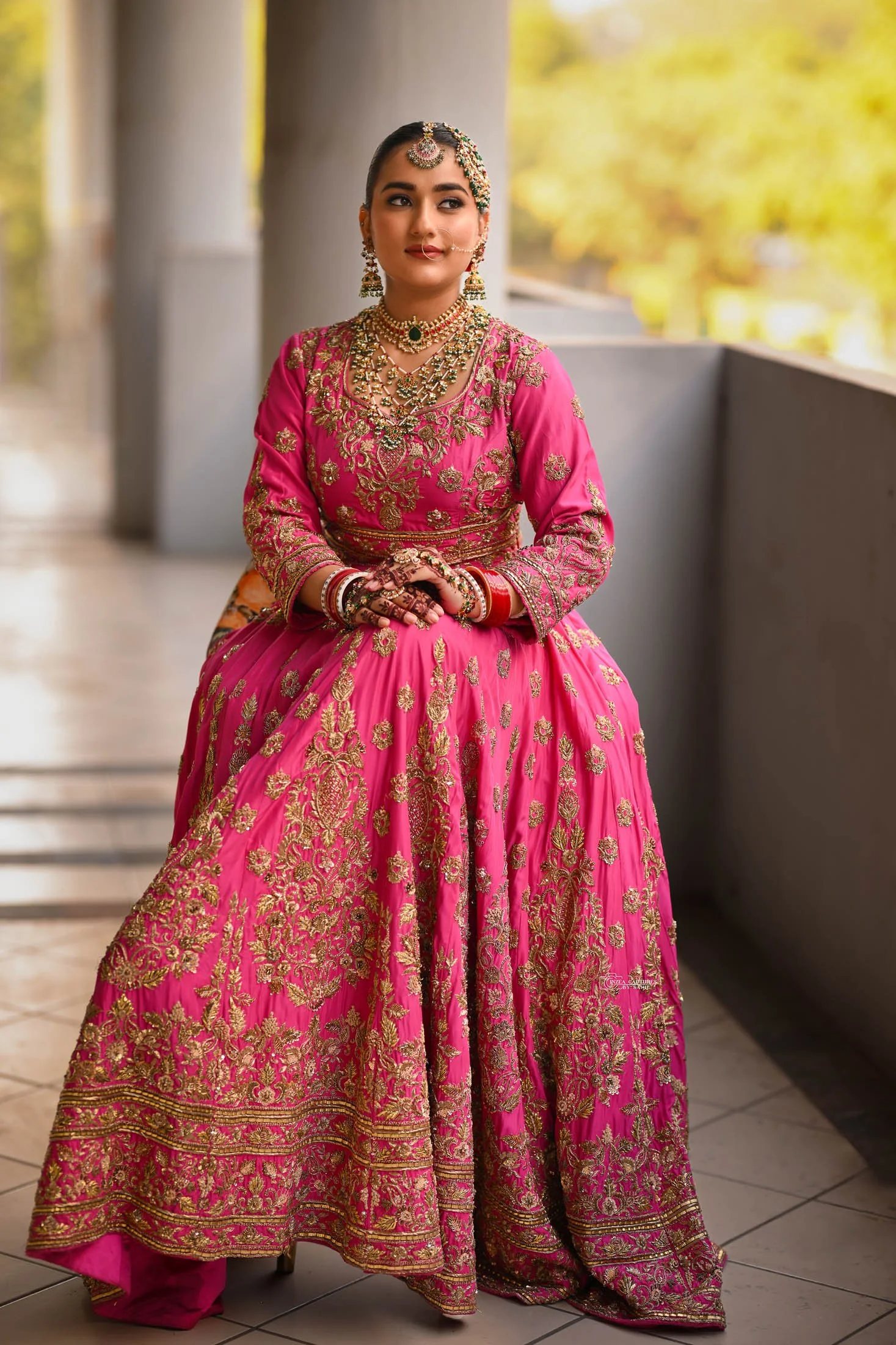 A woman dressed in a traditional pink and gold embroidered Indian bridal outfit, adorned with elaborate jewelry, sitting on a balcony with a background of blurred trees and sunlight.