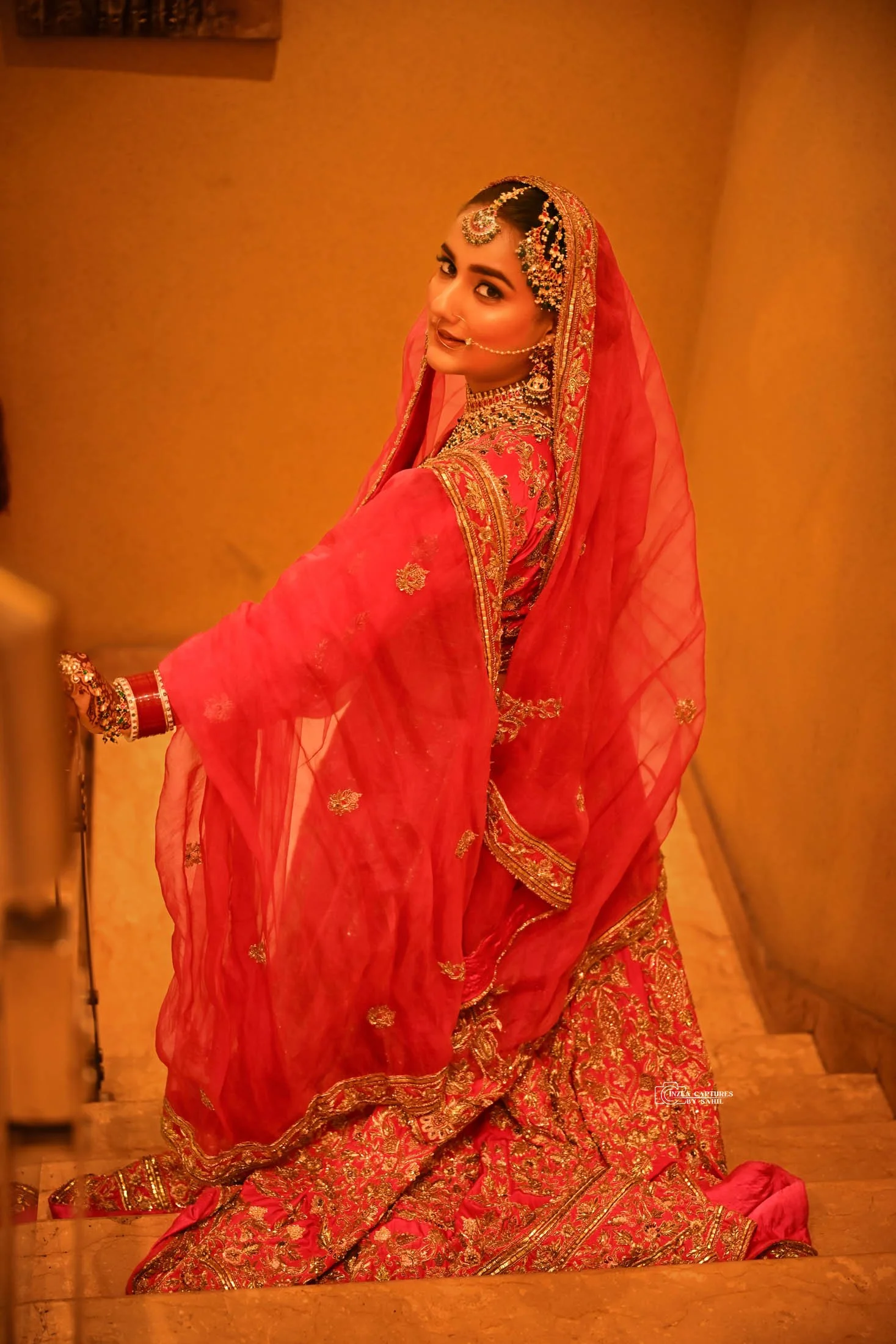 A woman dressed in traditional Indian bridal attire, wearing a red embroidered saree and ornate jewelry, standing on a staircase with a warm, amber-toned background.