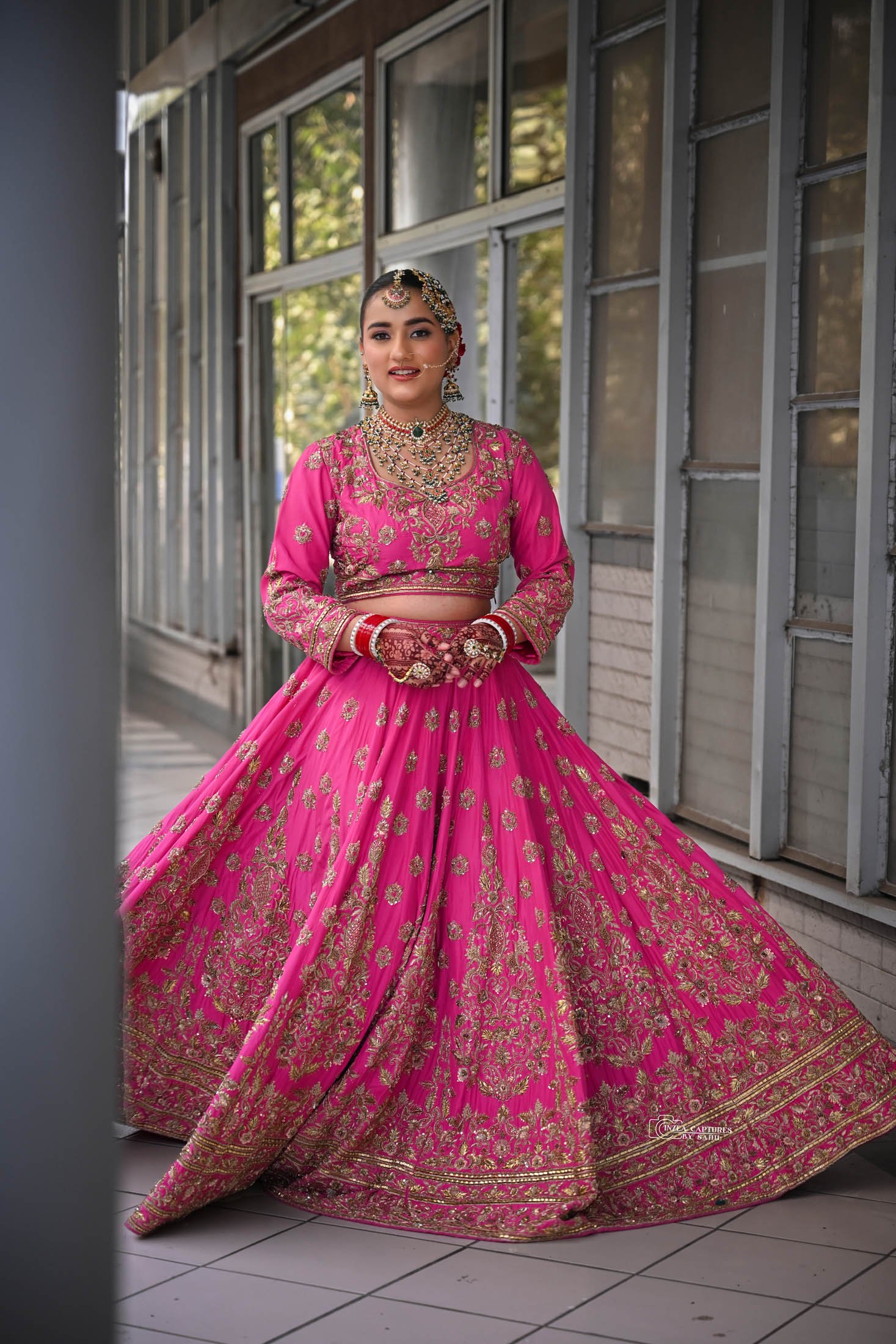 A woman dressed in a vibrant pink traditional Indian outfit with gold embroidery, adorned with jewelry including necklaces, earrings, bangles, and a headpiece, standing indoors near a large window with a background of trees.