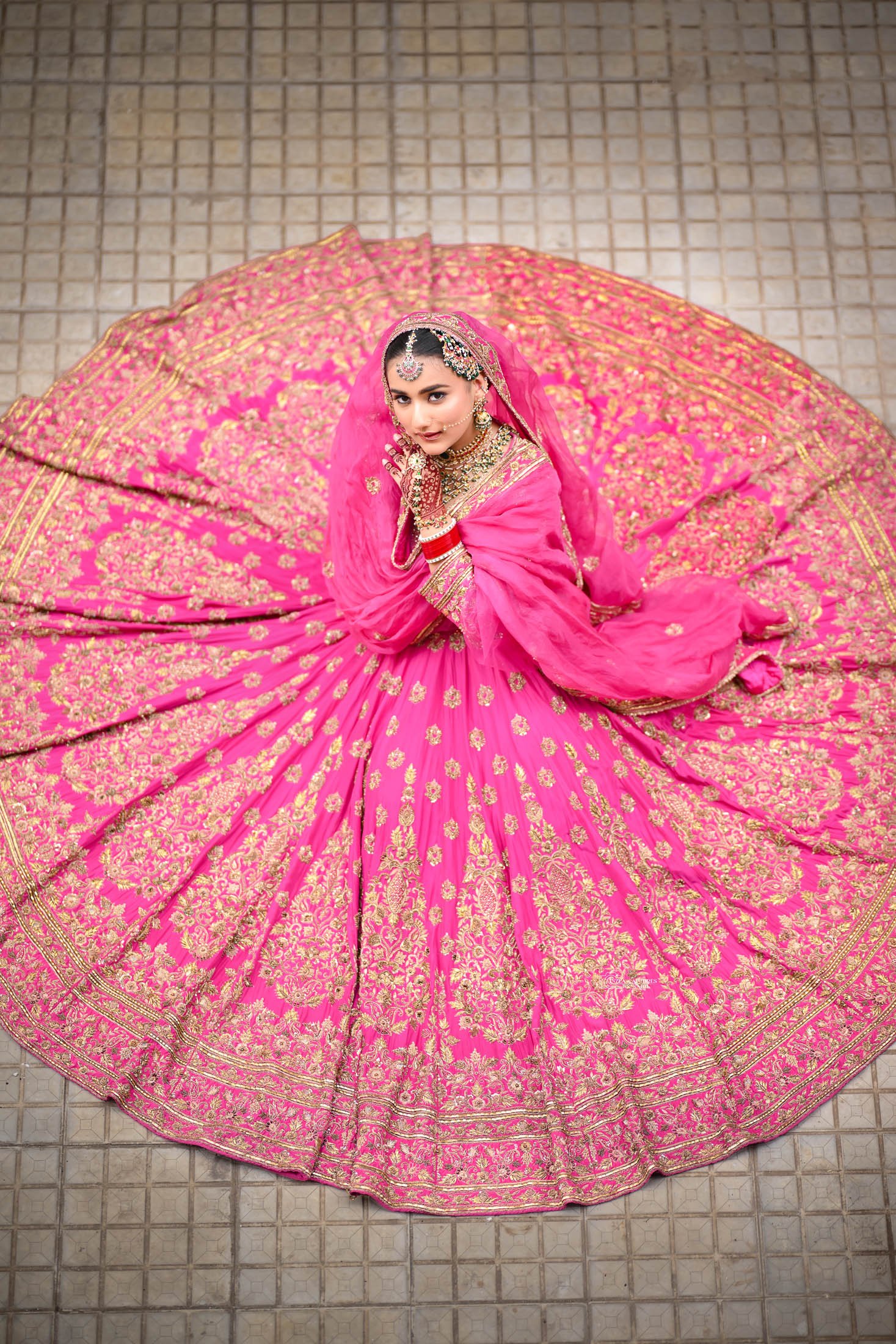 A woman in traditional Indian bridal attire sitting on a tiled floor, looking up at the camera, wearing a vibrant pink embellished lehenga with gold embroidery, jewelry, and a head covering.