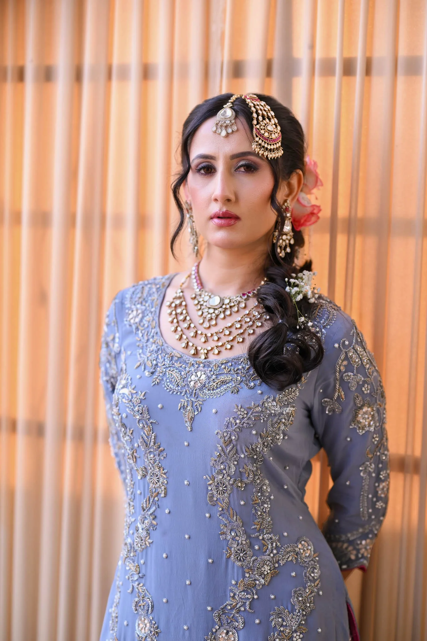 A woman dressed in traditional South Asian attire, adorned with intricate jewelry, standing in front of a beige curtain.