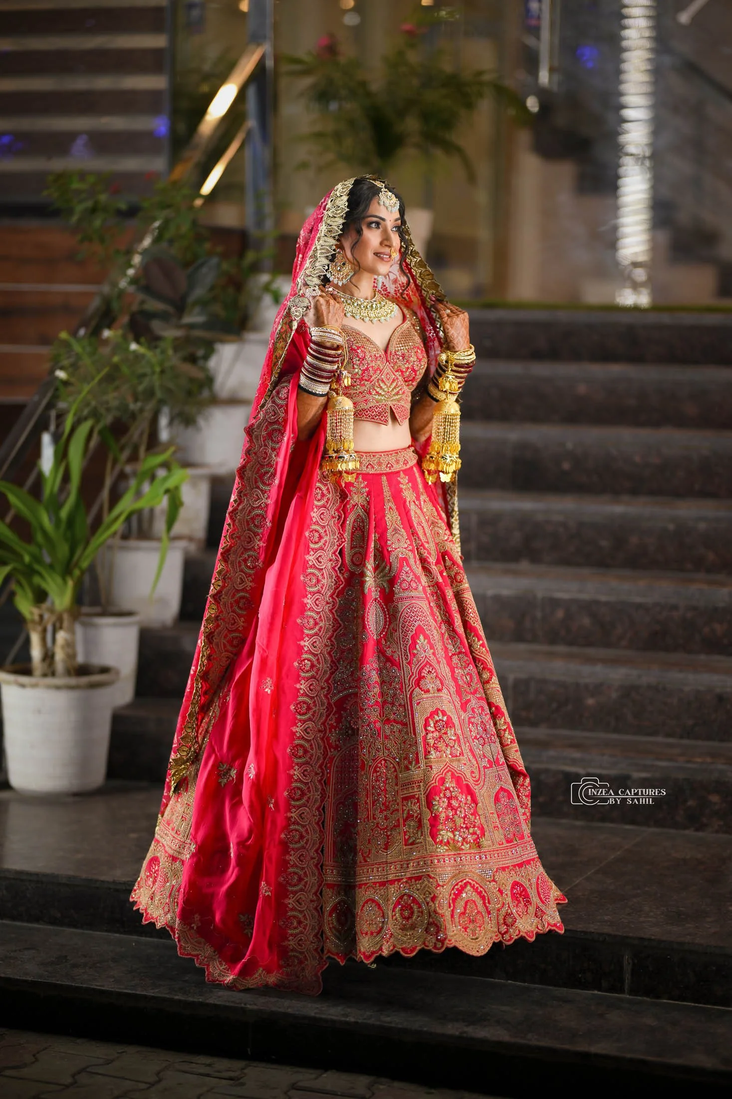 A woman dressed in traditional Indian wedding attire, including a red embroidered lehenga, dupatta, and gold jewelry, standing on a staircase.