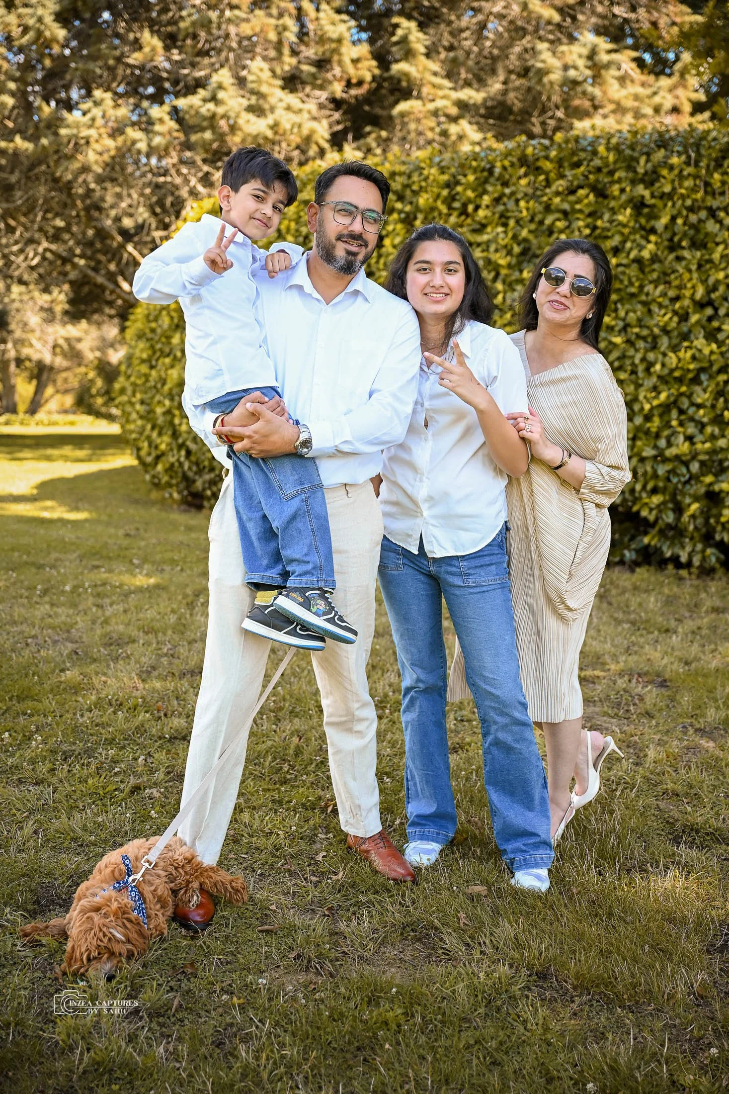 A family of four, including a young boy, a man, a woman, and an older woman, standing outdoors on a grassy area with trees and bushes in the background. The man is holding the boy. A small brown dog on a leash is in the foreground.