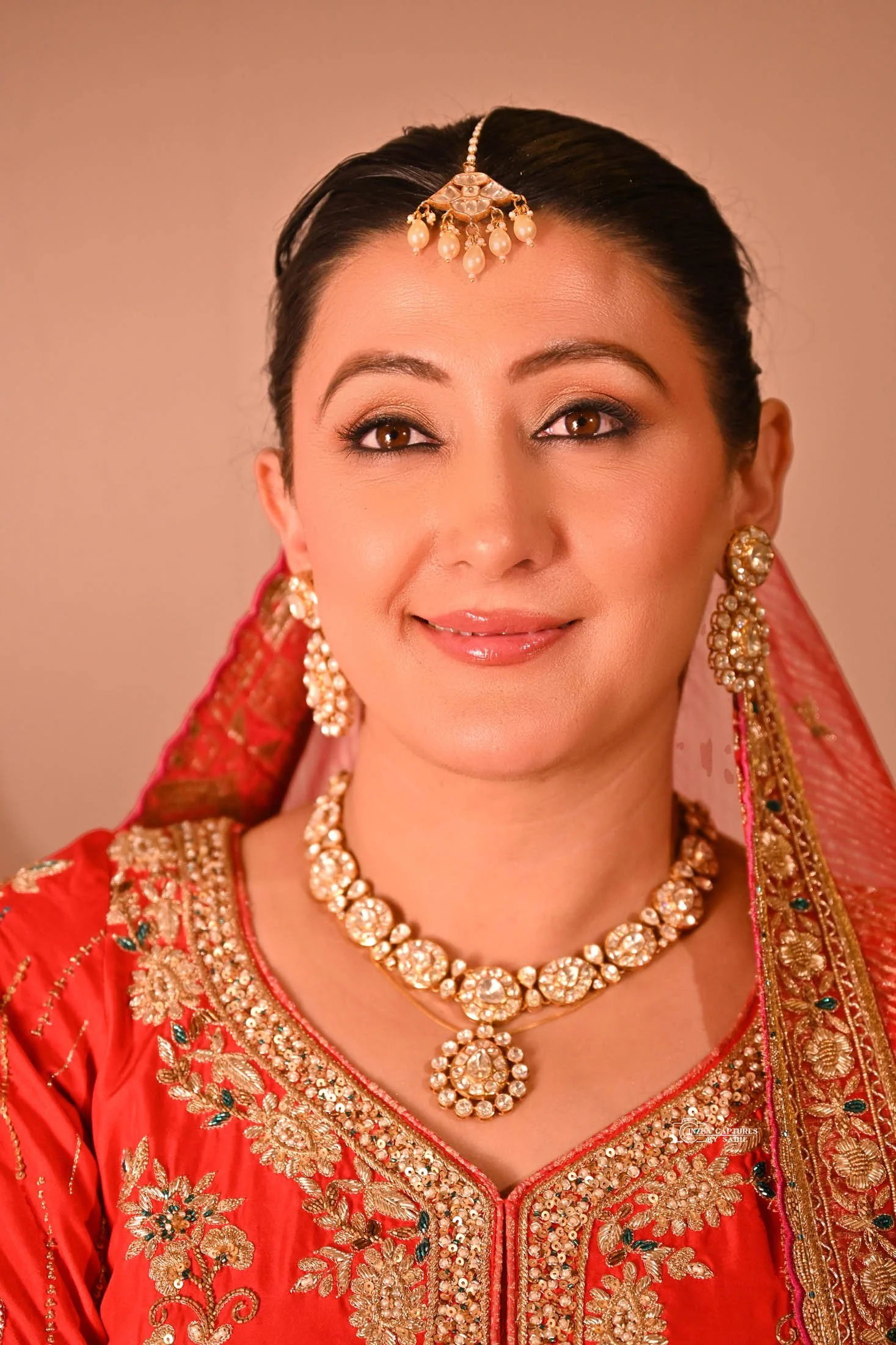 A woman dressed in traditional Indian clothing and jewelry, wearing a red embroidered outfit with gold accents, a matching red dupatta, and elaborate gold and pearl jewelry including earrings, necklace, and headpiece.