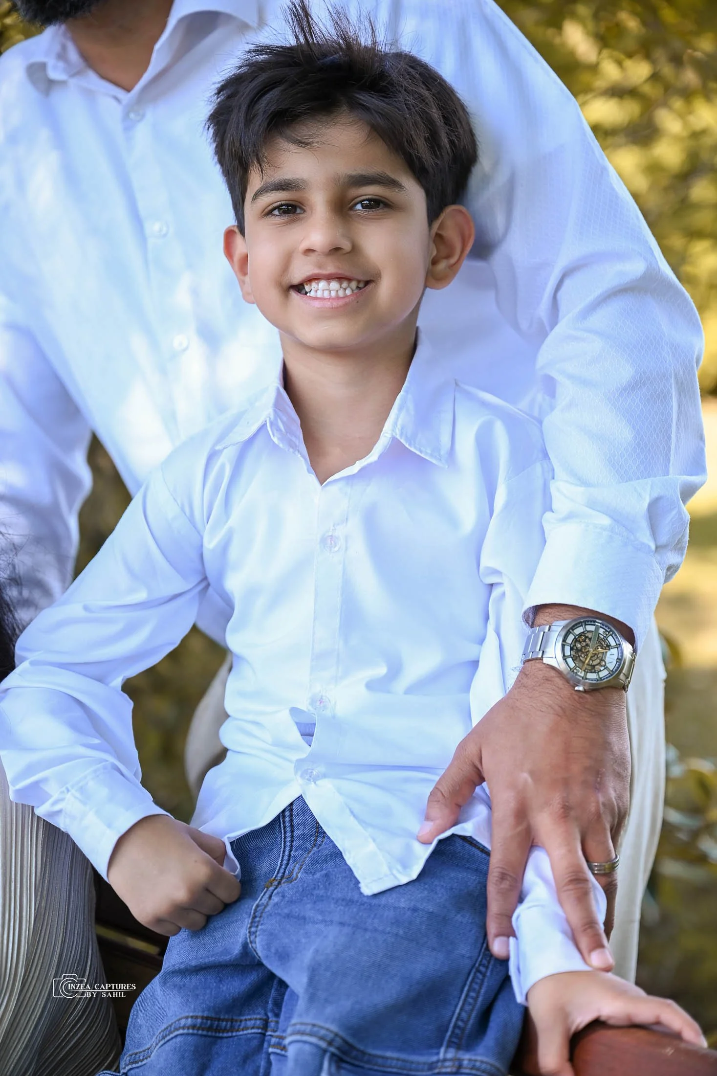 A smiling boy with dark hair wearing a white button-down shirt and jeans, sitting outdoors with a person behind him in a white shirt and an outdoor setting with trees in the background.
