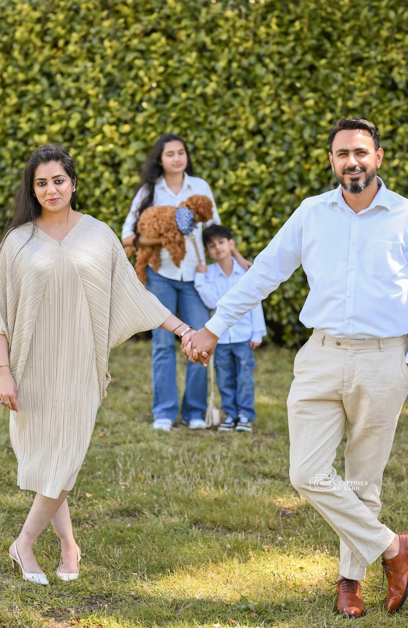 A happy family holding hands outdoors, with two children and a woman holding a small brown dog, against a background of green foliage.