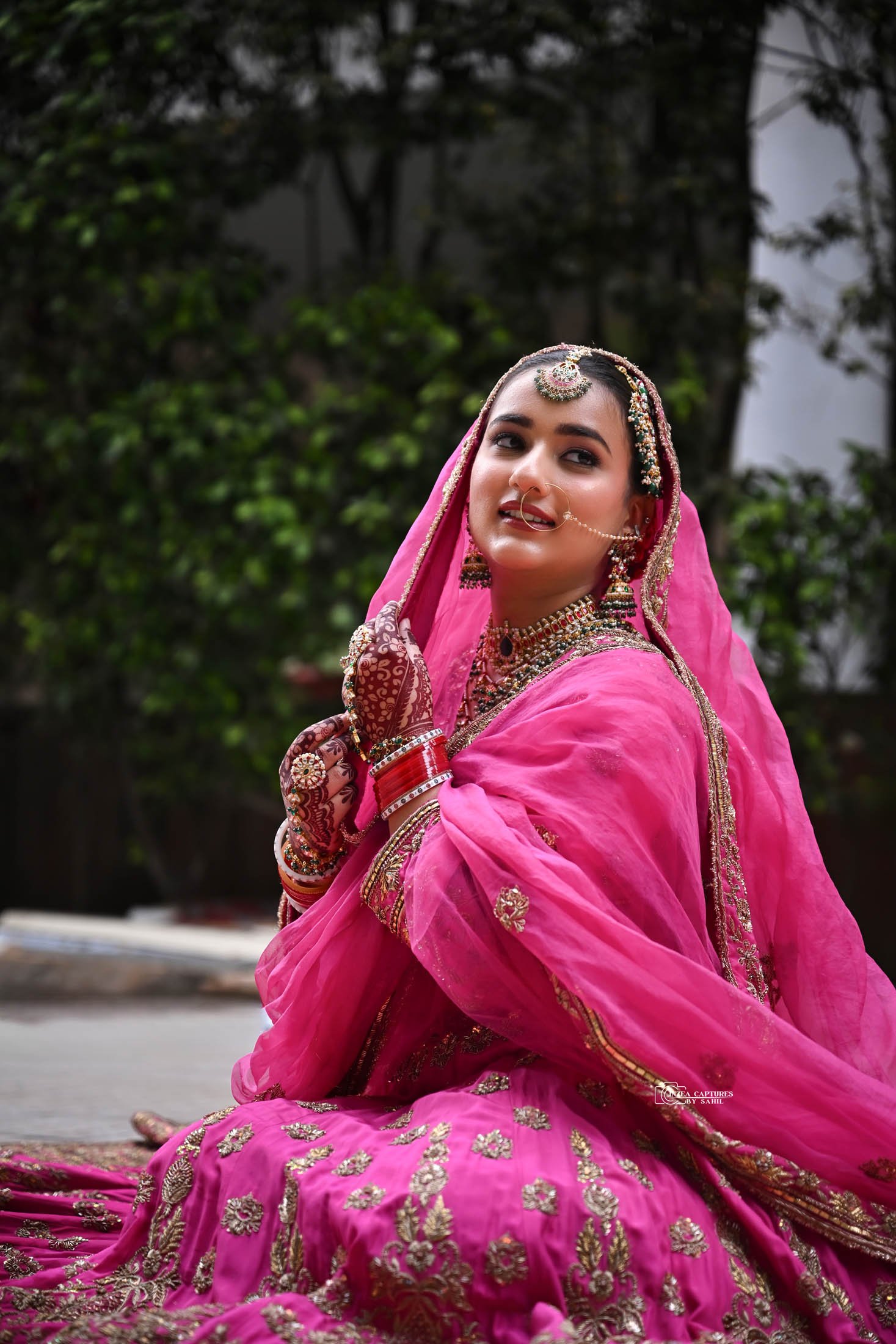 A woman dressed in traditional Indian attire, wearing a pink saree with gold embroidery, jewelry including a necklace, earrings, nose ring, and bangles, with henna on her hands, sitting outdoors with a green leafy background.