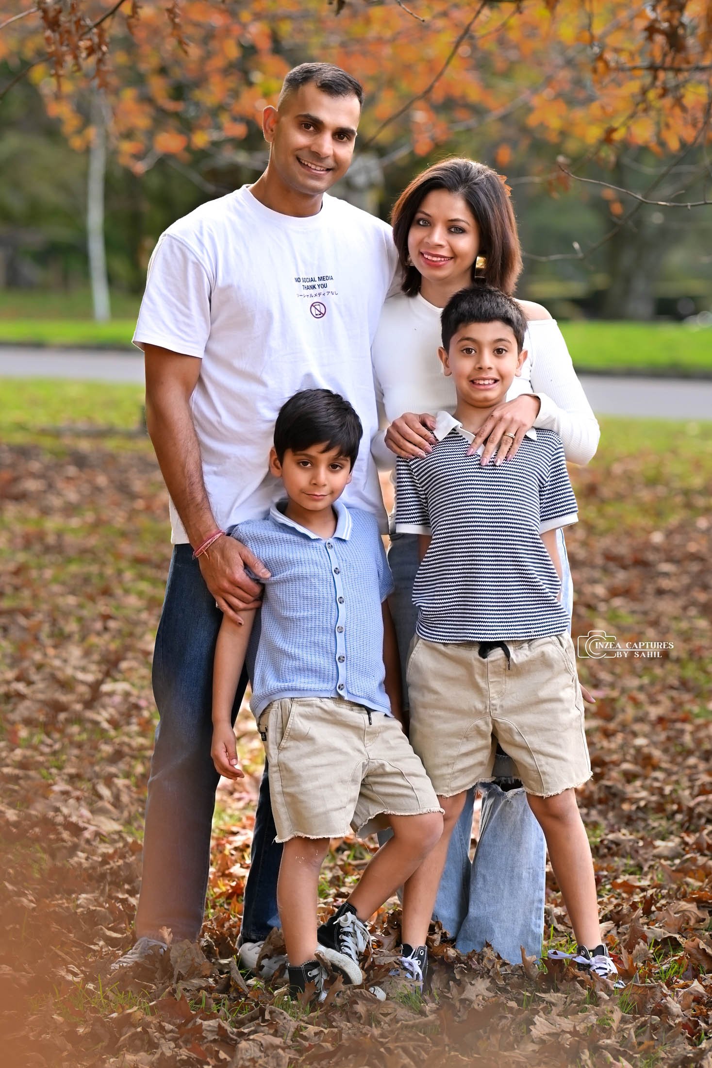 A family of five enjoying fall outdoors, standing on fallen leaves, with trees showing autumn colors in the background.