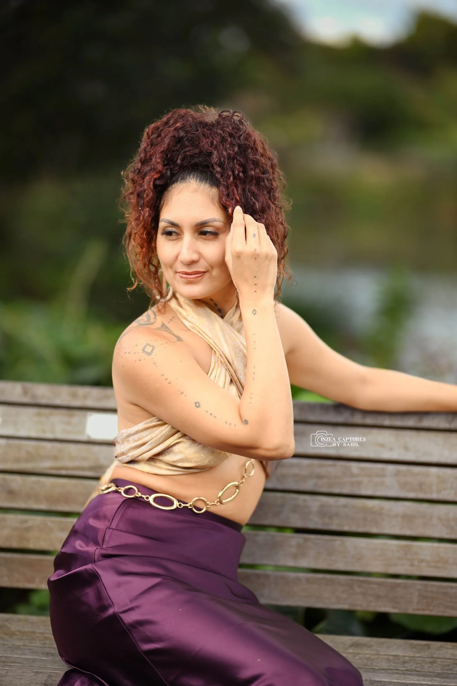 A woman with curly red hair sitting on a wooden bench outdoors, wearing a gold top, purple satin skirt, and a gold chain belt, with tattoos visible on her arm and shoulder, looking to the side with a gentle expression.
