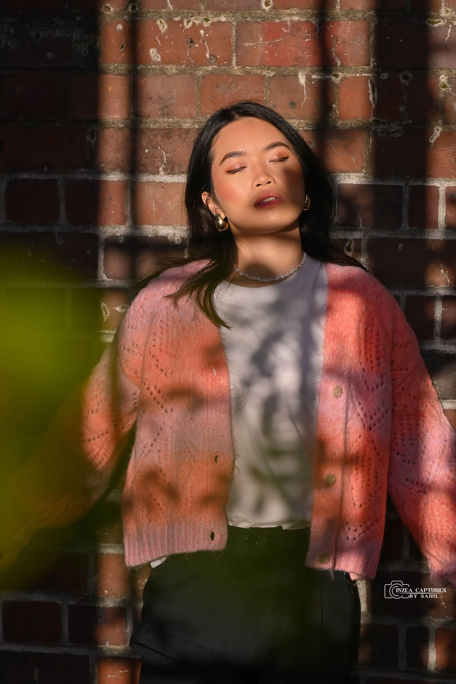 Woman with dark hair and earrings relaxing with eyes closed against a brick wall, sunlight creating shadows, wearing a pink cardigan, white top, and accessories.