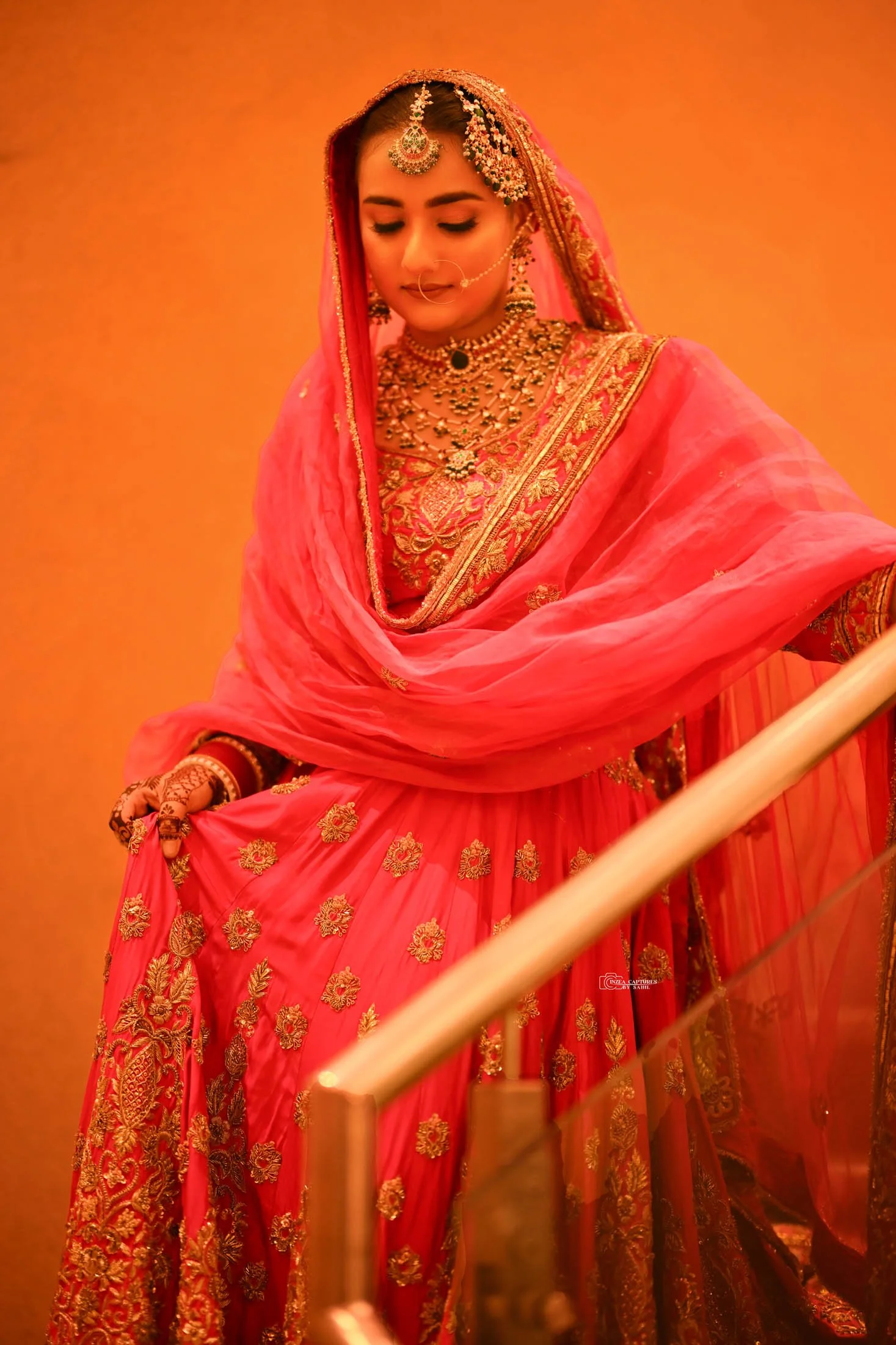 A woman wearing a pink traditional Indian bridal outfit with gold embroidery, jewelry, and a nose ring, standing against an orange background.